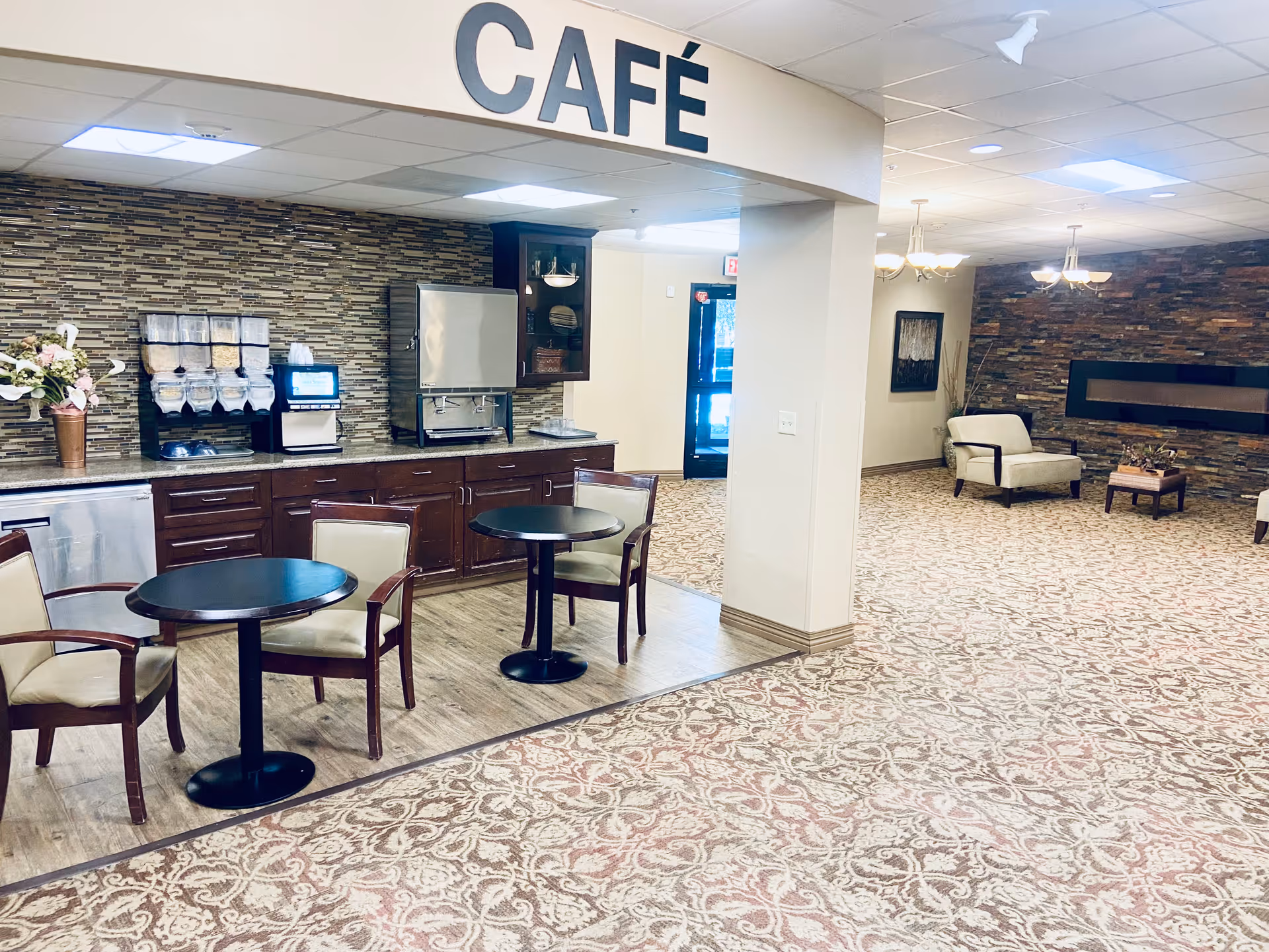 Interior view of a café area in a senior living facility with two small round tables and chairs, a counter with beverage dispensers and a microwave, and a seating area with armchairs and a stone accent wall with a modern fireplace.
