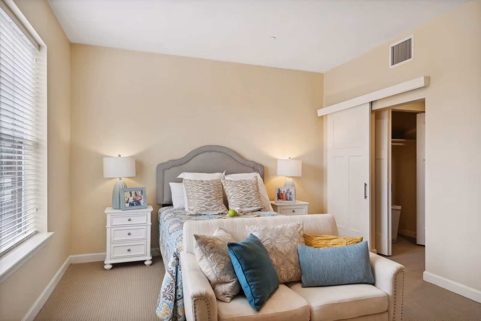 A cozy bedroom featuring a bed with a gray upholstered headboard, patterned pillows, and a colorful quilt. There are two white nightstands on either side of the bed, each with a lamp and framed photos. In front of the bed is a small beige loveseat with multiple decorative pillows. A large window with blinds is on the left wall, and a sliding door closet is visible on the right side of the room.