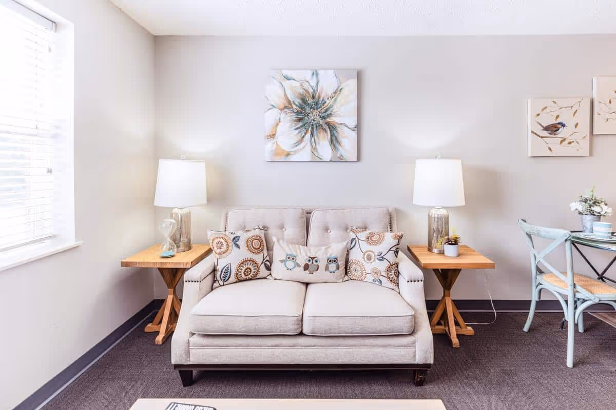 A cozy living room area with a beige loveseat adorned with decorative pillows featuring owl and floral designs. On either side of the loveseat are wooden side tables with lamps and small plants. The walls are decorated with floral and bird artwork, and a window with blinds allows natural light to brighten the space. A light blue chair and a small dining table are partially visible on the right side.