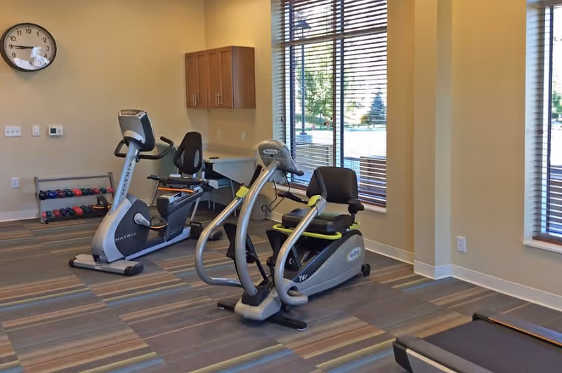 A small exercise room with two stationary recumbent exercise bikes, a rack of colorful dumbbells, a wall clock, and large windows with blinds letting in natural light.