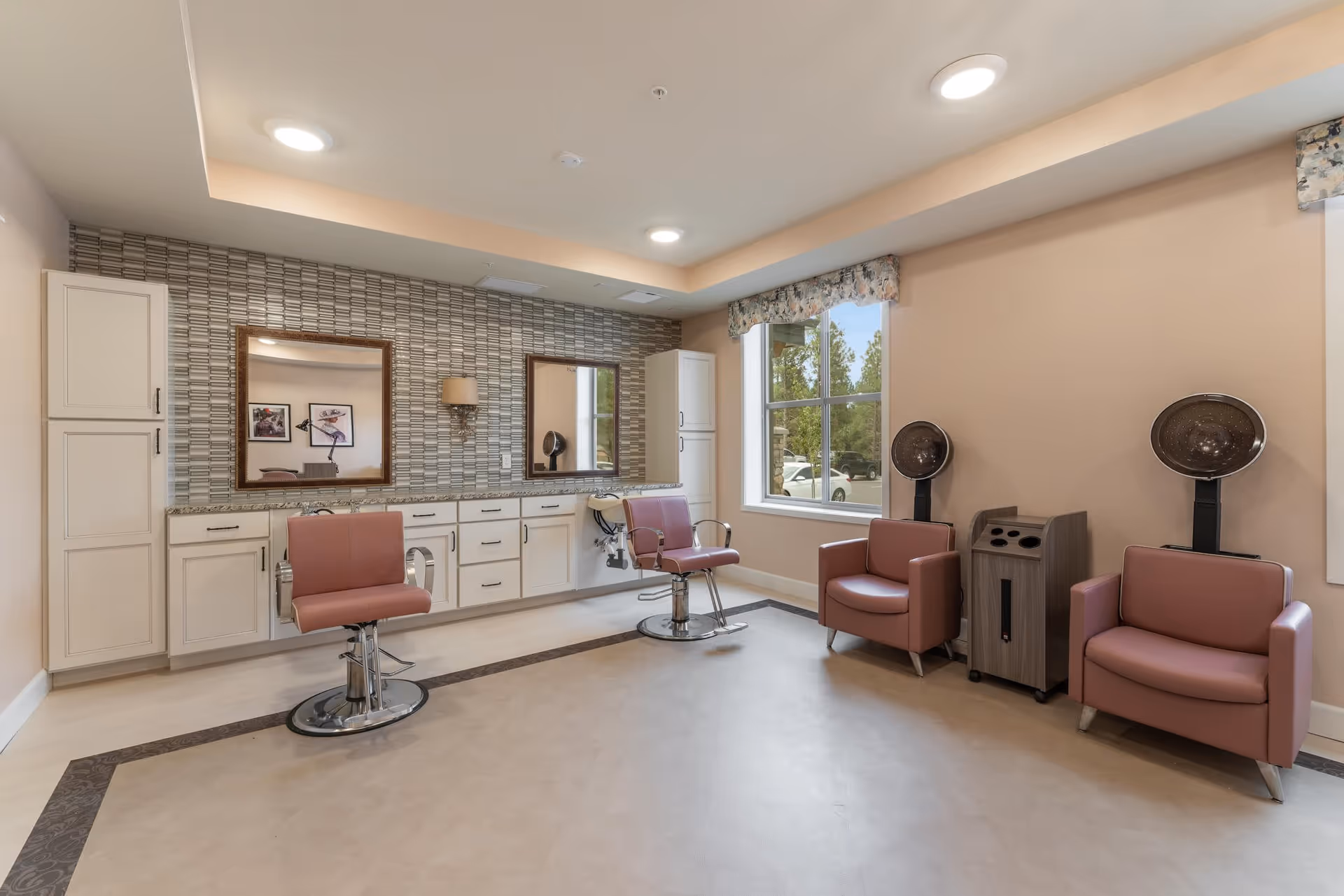 Interior view of a senior living facility's hair salon with two pink salon chairs in front of a counter with cabinets and two large mirrors. There are two pink armchairs with hair dryers next to a window showing a view of parked cars and trees outside.