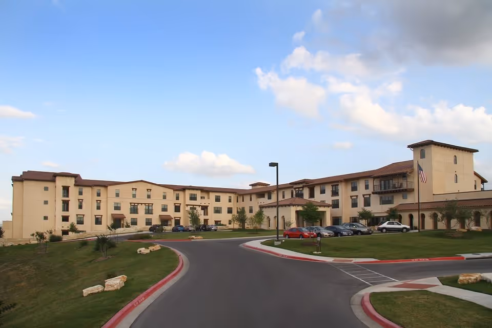 Exterior view of a large, three-story senior living facility building with beige walls and a red-tiled roof under a partly cloudy blue sky. There is a curved driveway in front with several parked cars and a grassy area with rocks and small trees.