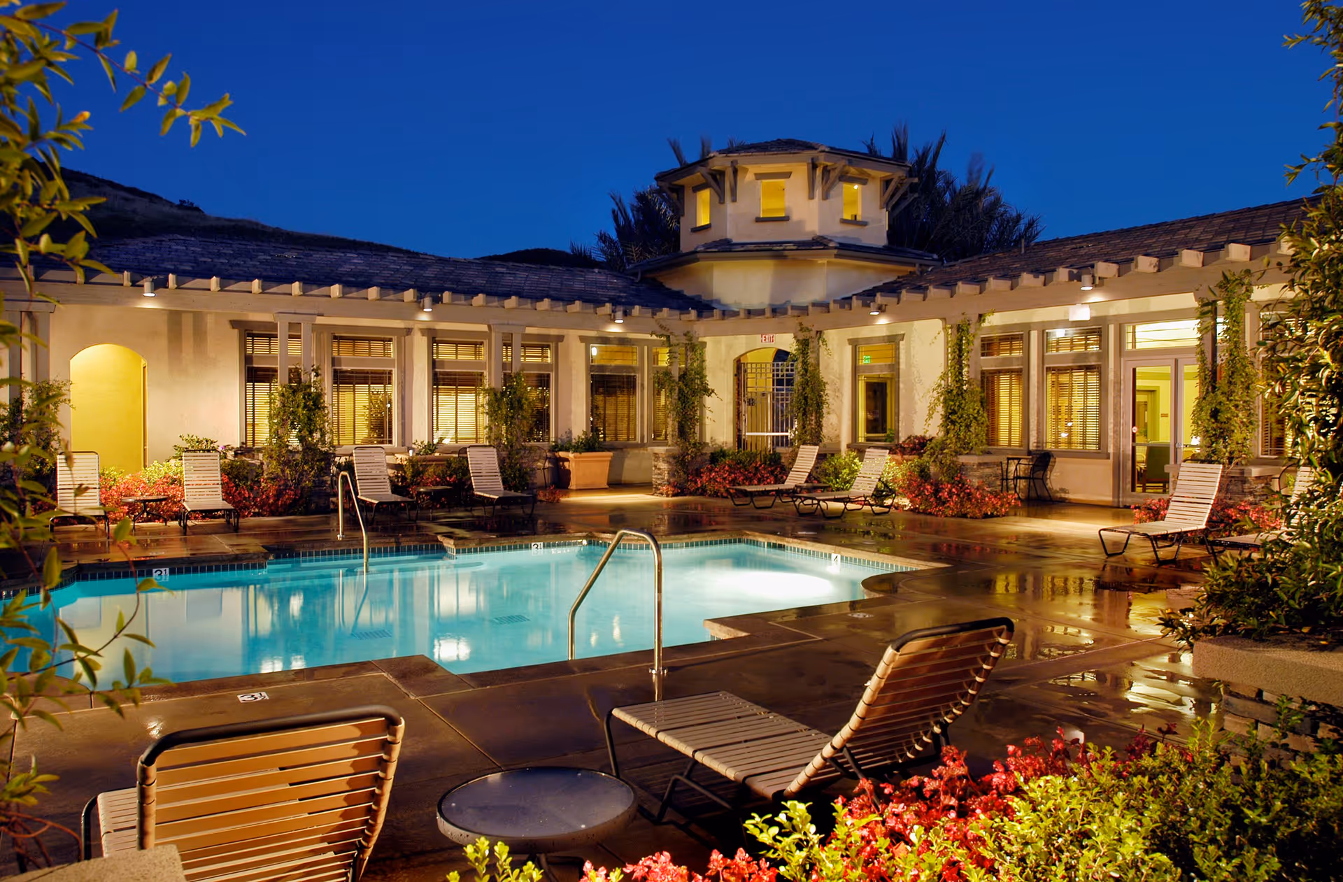 Illuminated outdoor swimming pool at dusk surrounded by lounge chairs and a courtyard-style building.