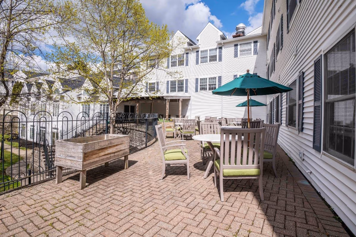 Outdoor patio area at Middlebrook Farms at Trumbull with several round tables and chairs with green cushions, green umbrellas providing shade, a wooden planter box, and a tree with budding leaves. The patio is paved with brick and surrounded by white buildings with multiple windows under a partly cloudy sky.