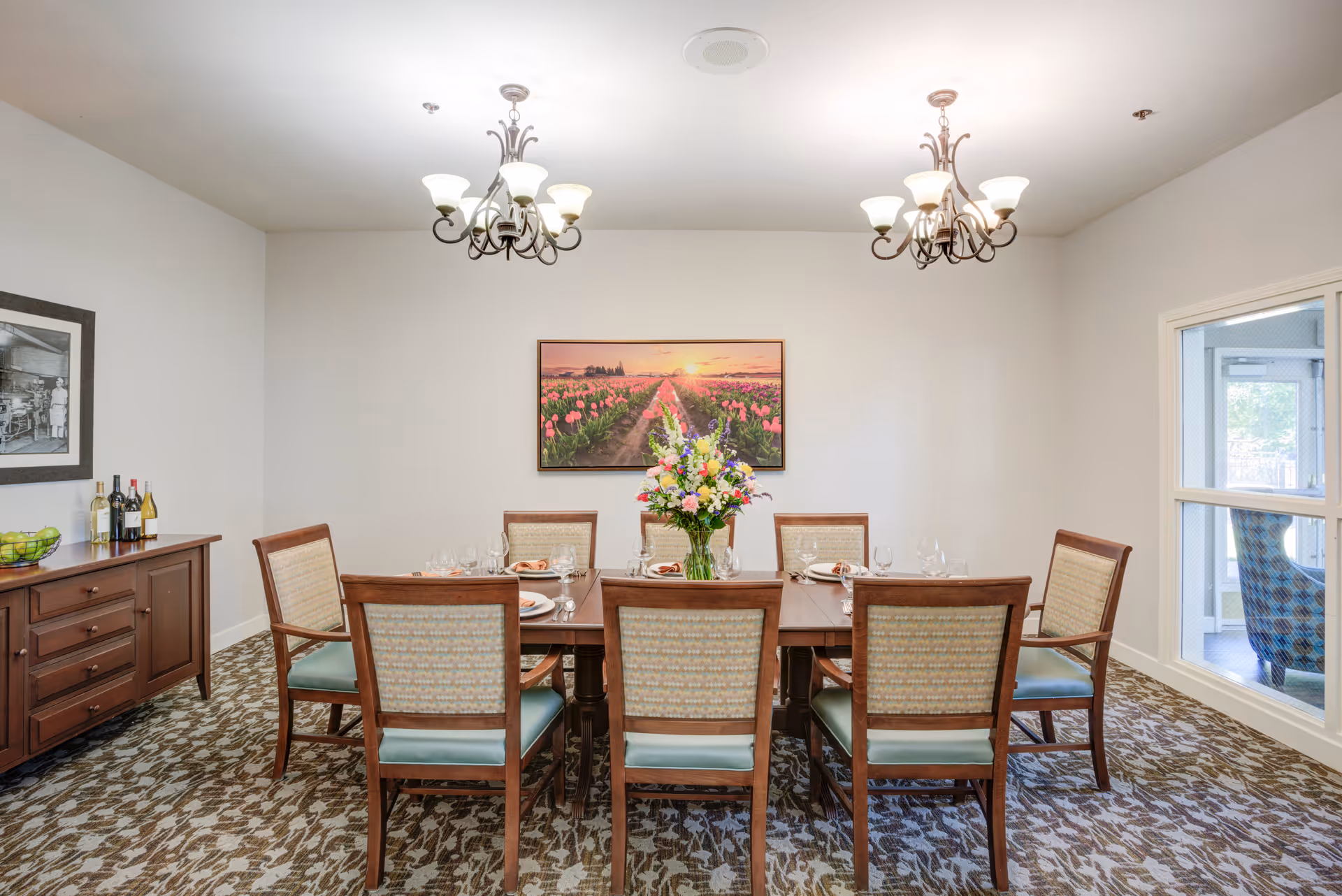 A dining room with a rectangular wooden table set for six people. There are six wooden chairs with cushioned seats around the table. A vase with a colorful flower arrangement is placed in the center of the table. On the left side, there is a wooden sideboard with bottles of wine and a bowl of green apples. The room has two chandeliers hanging from the ceiling and a framed picture of a tulip field at sunset on the wall. A window on the right side shows a glimpse of another room with a blue patterned chair.