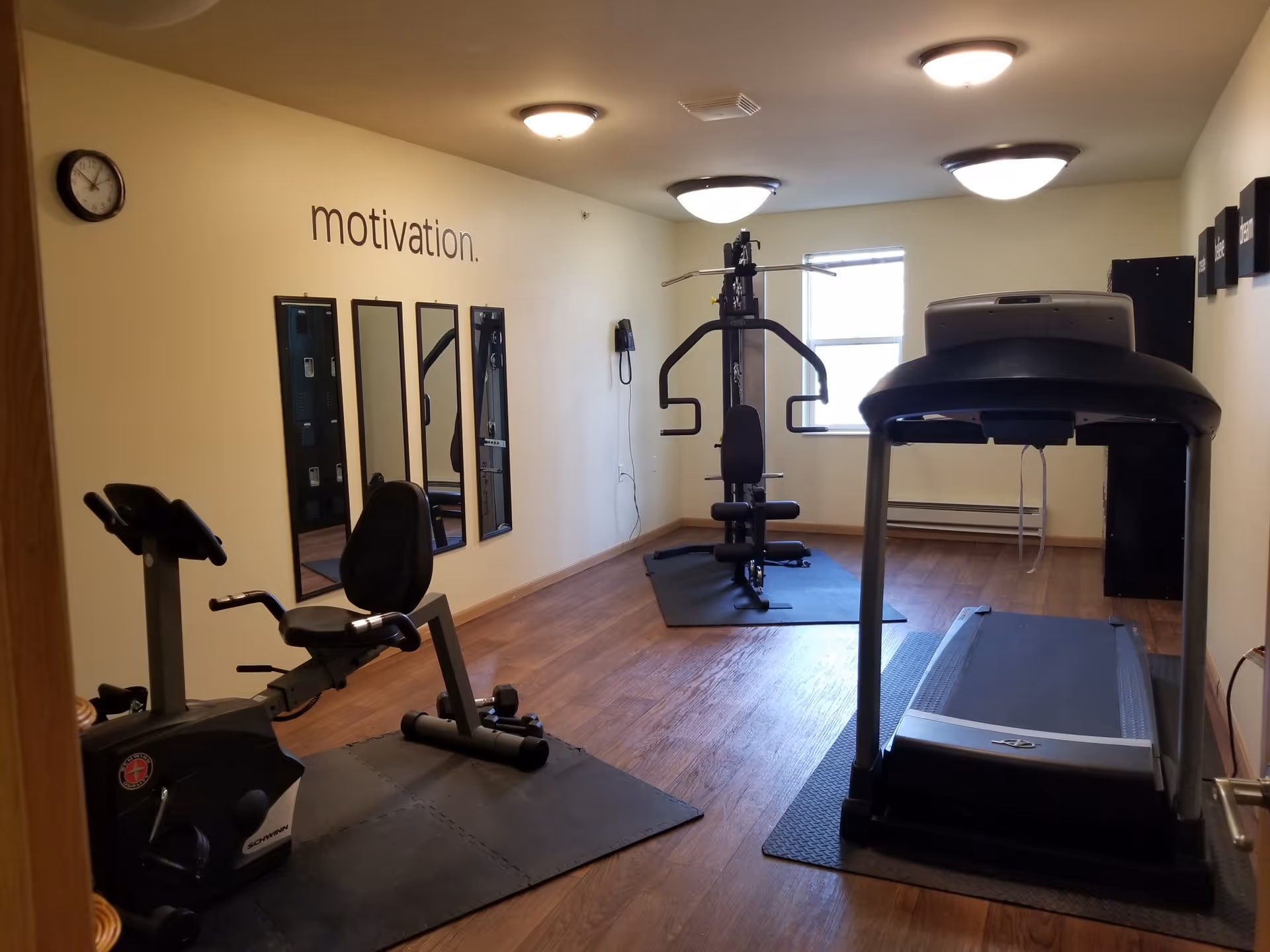 A small fitness room with exercise equipment including a recumbent bike, a multi-function weight machine, and a treadmill. The room has wooden flooring, three ceiling lights, a wall clock, three vertical mirrors, and the word 'motivation.' written on the wall. There is a window letting in natural light and a telephone mounted on the wall.