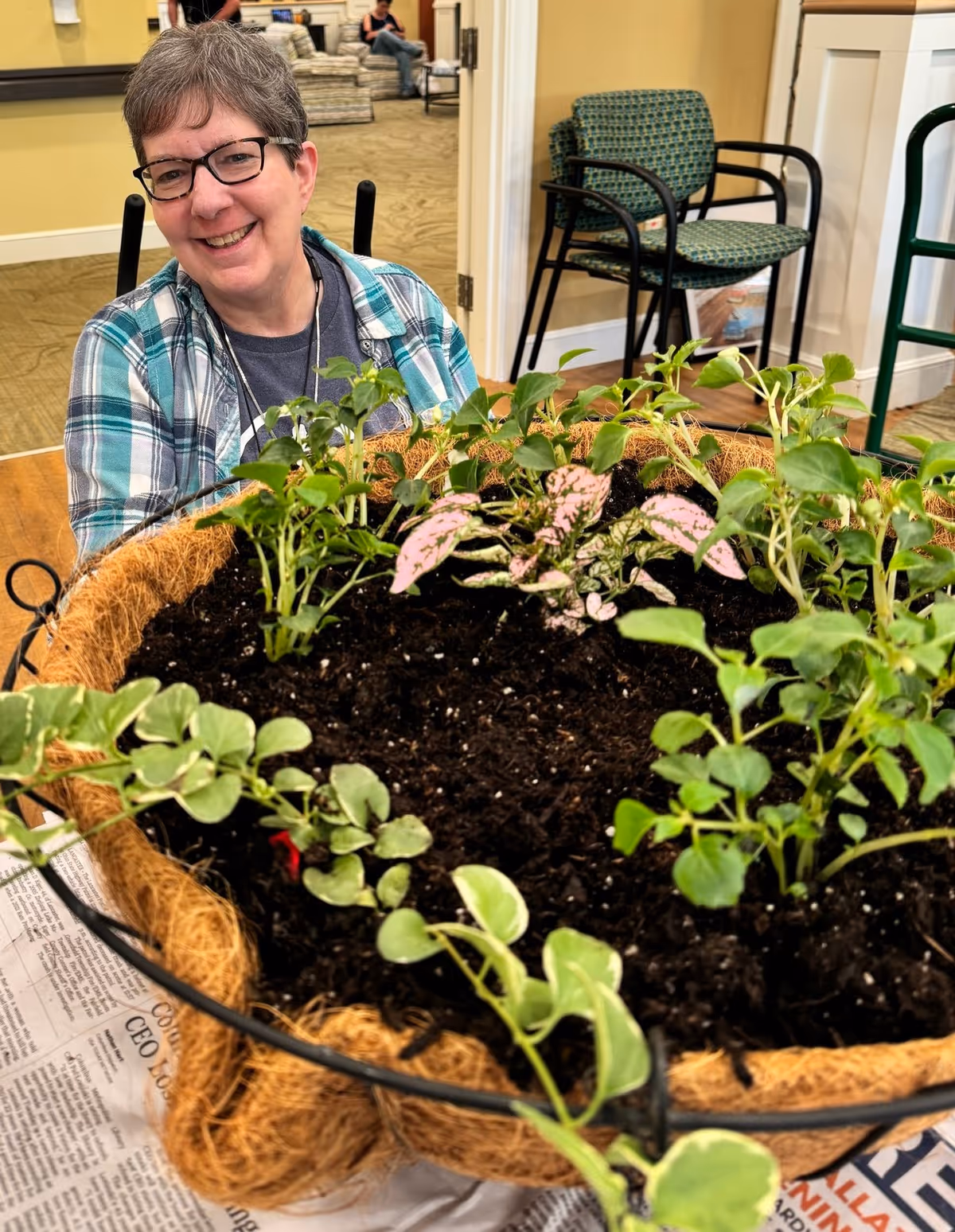 A smiling woman with short gray hair and glasses is sitting indoors behind a large planter filled with soil and various small green plants. The room has a carpeted floor, a couple of chairs, and a person sitting in the background.