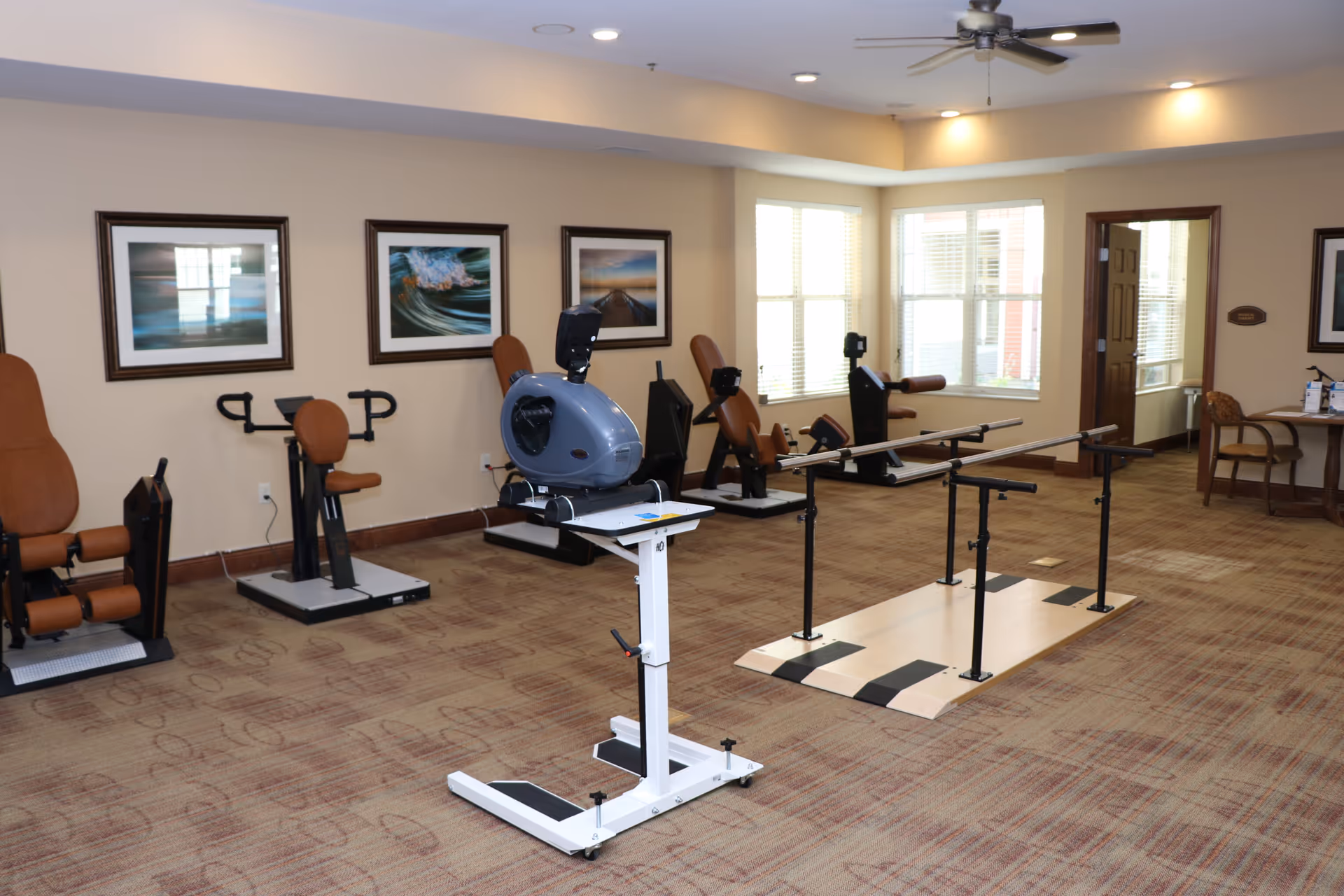 A senior living facility exercise room with various exercise equipment including a stationary bike, leg press machines, and parallel bars for walking support. The room has beige walls, carpeted floor, framed pictures on the wall, large windows letting in natural light, and a ceiling fan.