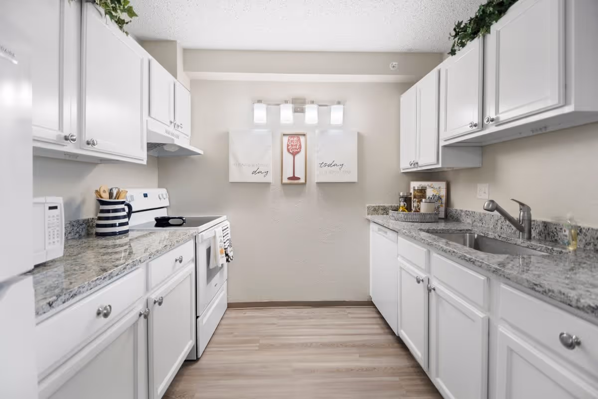 A clean, modern kitchen with white cabinets and granite countertops on both sides. The kitchen features a white stove with a microwave above it on the left side and a stainless steel sink on the right side. Above the sink and stove are white upper cabinets. The floor is light wood, and there are three decorative wall hangings and a light fixture on the far wall. Some kitchen utensils and containers are placed on the countertops.