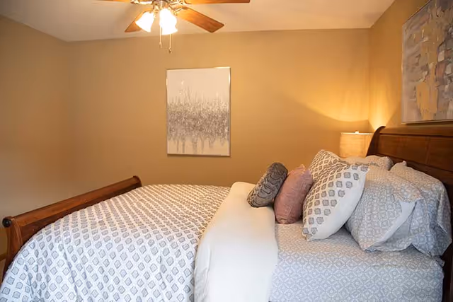A cozy bedroom with a wooden sleigh bed featuring patterned bedding and multiple pillows. The walls are painted beige, decorated with two abstract paintings. A ceiling fan with lights is mounted on the ceiling, and a bedside lamp provides warm lighting.