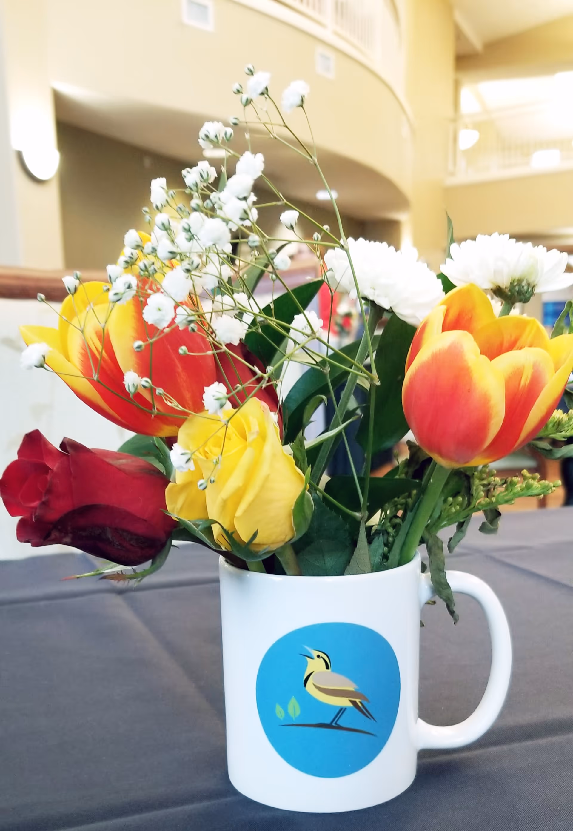 A white mug with a bird logo filled with a colorful bouquet of flowers including red and yellow tulips, white daisies, and baby's breath, placed on a table with a dark tablecloth in a bright indoor setting with a balcony and railing in the background.