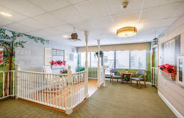 Bright senior living common area with a white decorative railing, seating by large windows, and floral wall murals.