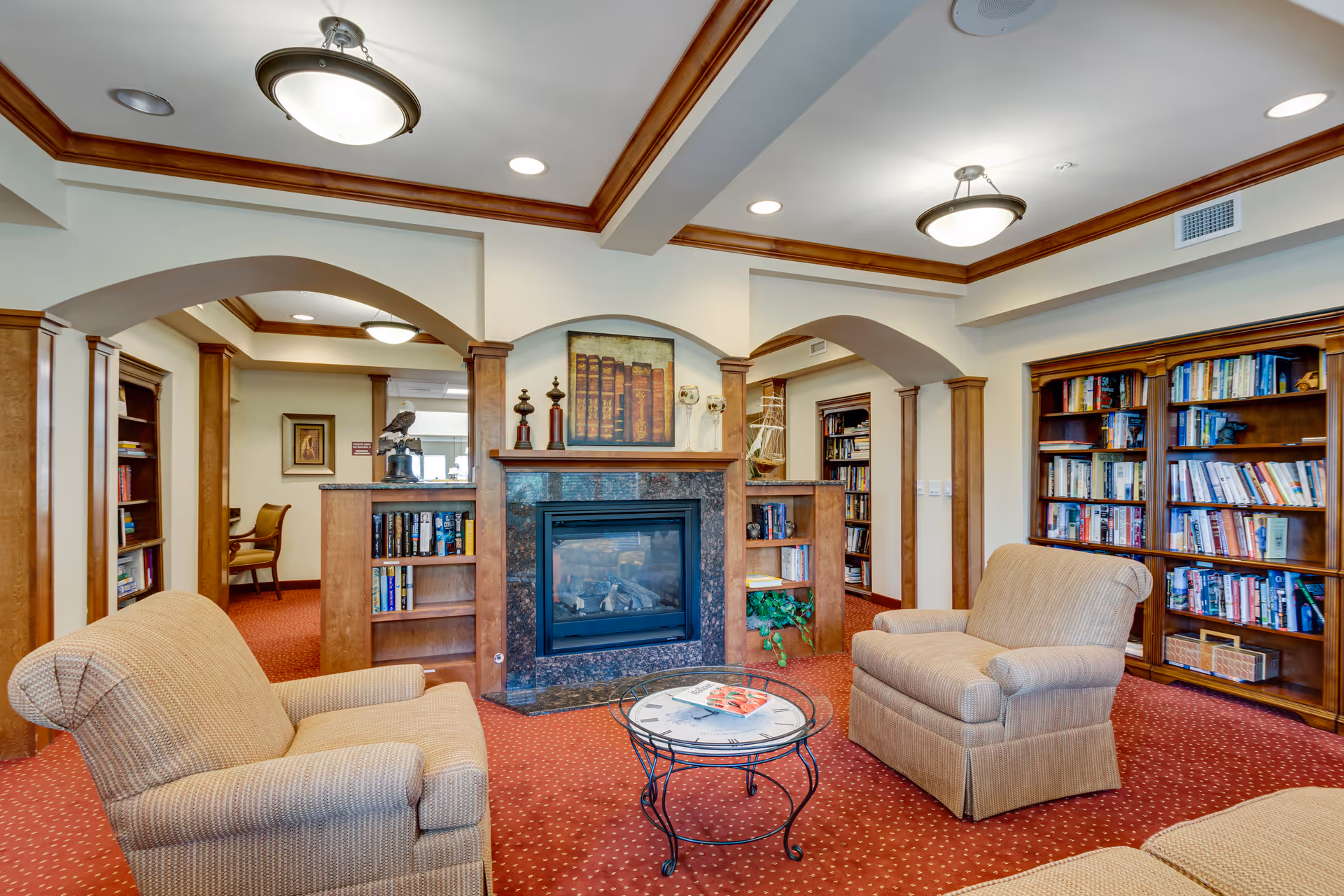 A cozy living room area in Magnolia Heights Gracious Retirement Living featuring two beige upholstered armchairs facing a glass-top round coffee table. The room has a fireplace with a stone surround and wooden mantle, flanked by built-in bookshelves filled with books. The ceiling has recessed lighting and decorative wooden trim. The carpet is red with a subtle pattern, and there are arched doorways leading to other rooms with more bookshelves and seating.