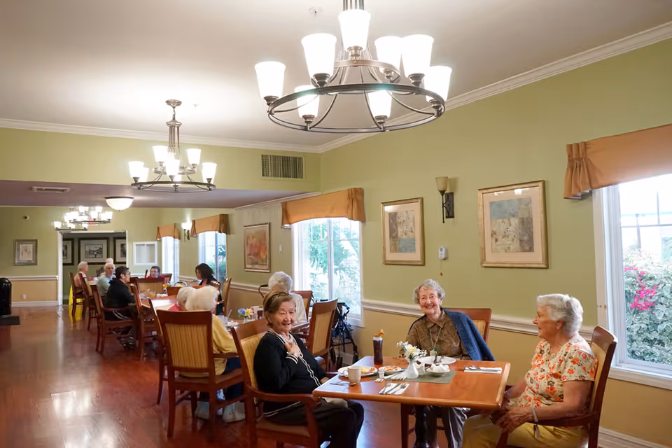 A dining room in a senior living facility with several elderly residents sitting at wooden tables, enjoying a meal and conversation. The room has green walls, large windows with orange valances, framed artwork, and multiple ceiling light fixtures.