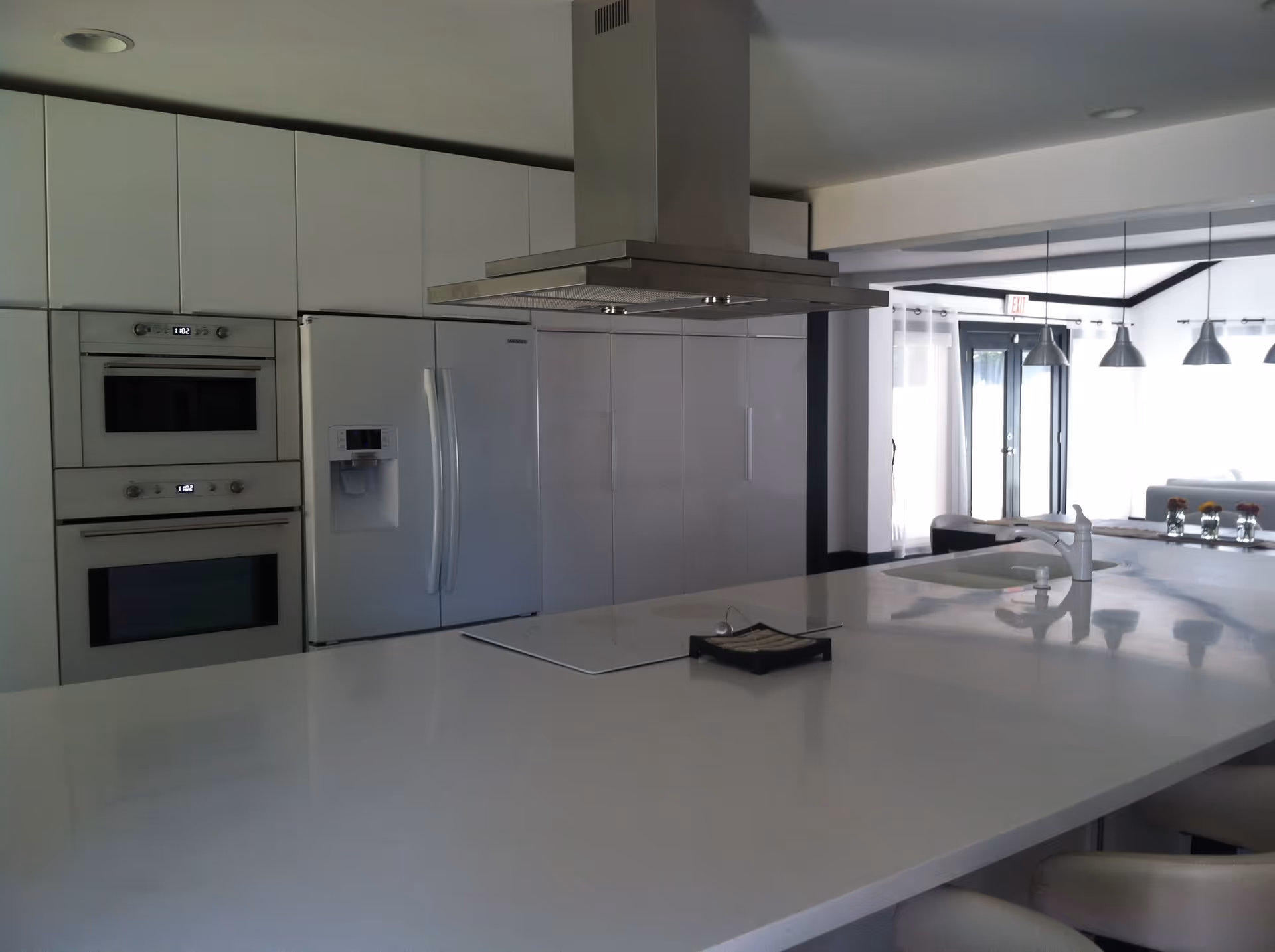 Modern kitchen with white cabinetry, a large white island countertop with a built-in sink and faucet, a stainless steel range hood above the island, double wall ovens, and a refrigerator with a water and ice dispenser. In the background, there is a dining area with hanging pendant lights and glass doors leading outside.