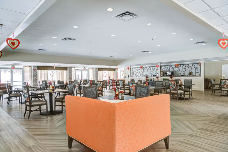 Large bright dining room with multiple tables and chairs and an orange corner banquette in the foreground.
