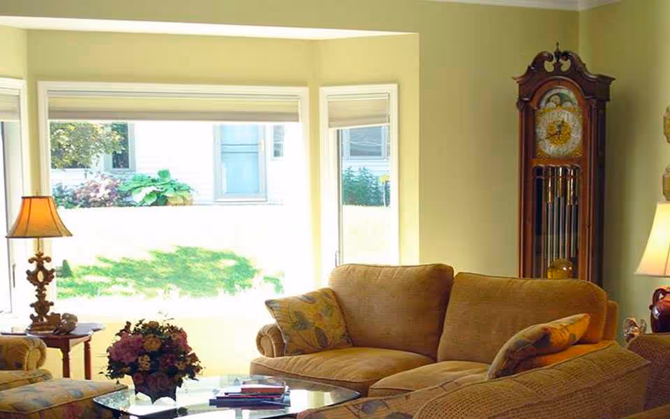 A cozy living room with beige sofas adorned with patterned cushions, a glass coffee table with a flower arrangement and books, a wooden grandfather clock, and two table lamps near large windows letting in natural light.