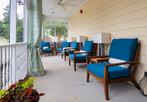 A covered outdoor porch area with several wooden chairs featuring blue cushions and white cylindrical pillows. The porch has a white railing, green curtains tied back, ceiling fans, and lamps placed on small tables between the chairs. There are plants visible in the foreground and greenery outside the porch.