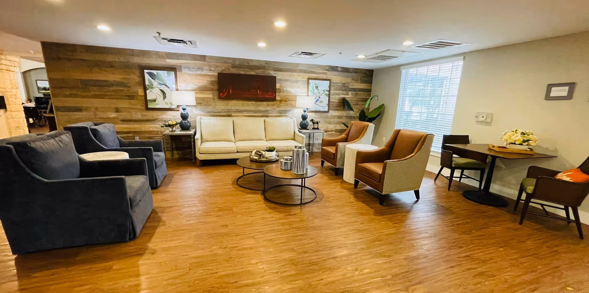 Spacious communal lounge with sofas, armchairs, round coffee tables and a wood-accent wall under recessed lighting.