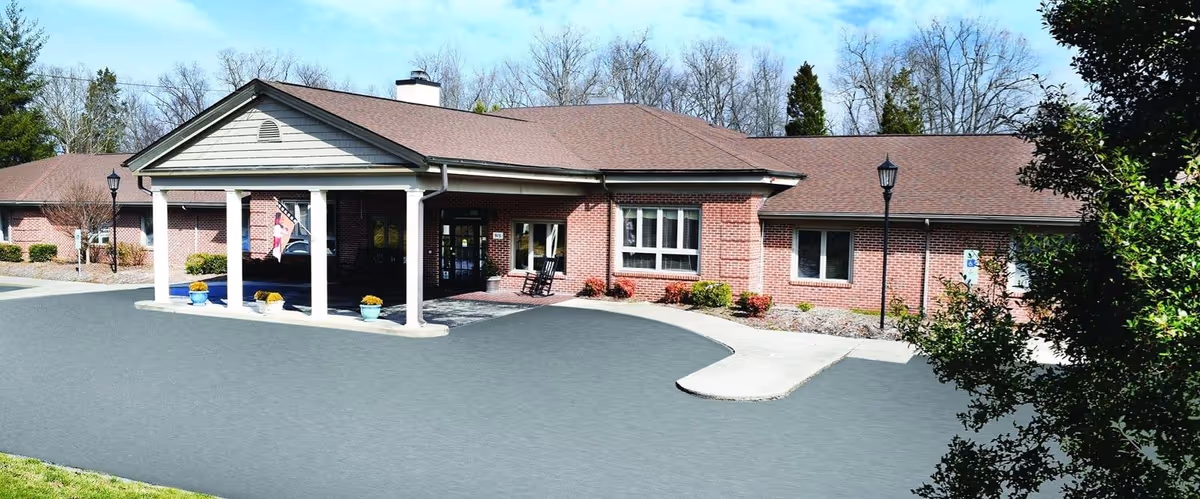 Exterior view of Brookstone Terrace of Thomasville Assisted Living and Memory Care building with a covered entrance supported by white columns, brick walls, and a driveway in front. There are some potted plants near the entrance and trees in the background.
