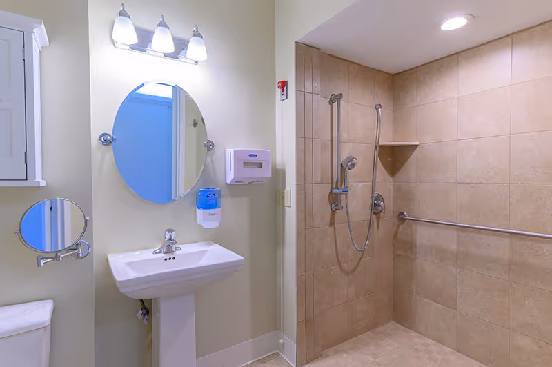 A clean and accessible bathroom featuring a pedestal sink with a round mirror above it, a wall-mounted soap dispenser and paper towel holder, a small adjustable round mirror, and a tiled walk-in shower with grab bars and a handheld showerhead.
