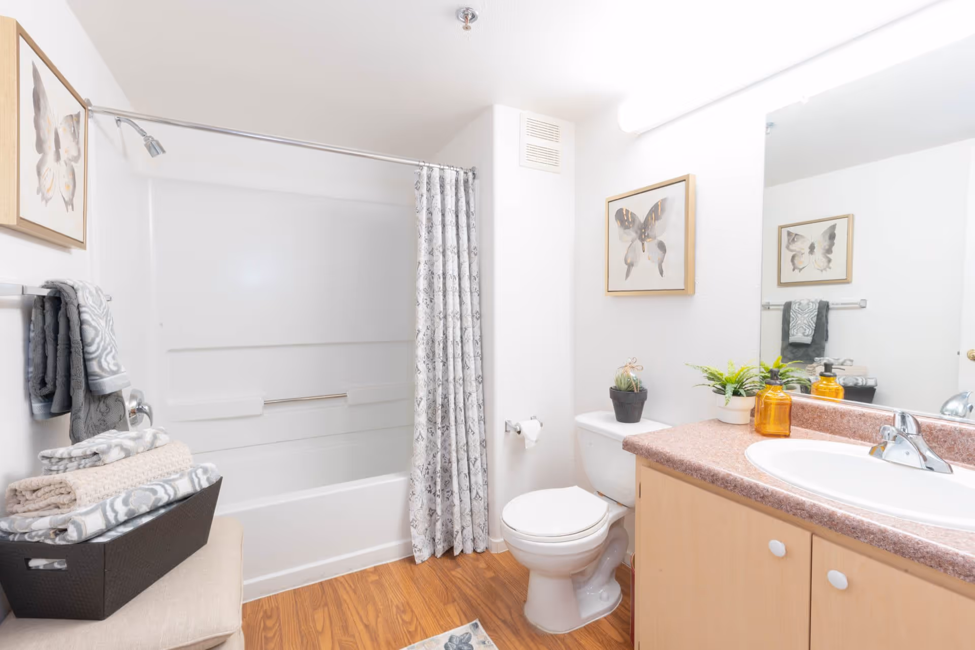 A clean bathroom with a white bathtub and shower combination, a patterned shower curtain, a toilet, and a vanity with a sink and countertop. There are framed butterfly pictures on the walls, a basket with folded towels on a bench, and small potted plants on the countertop. The floor has a wood-like finish.