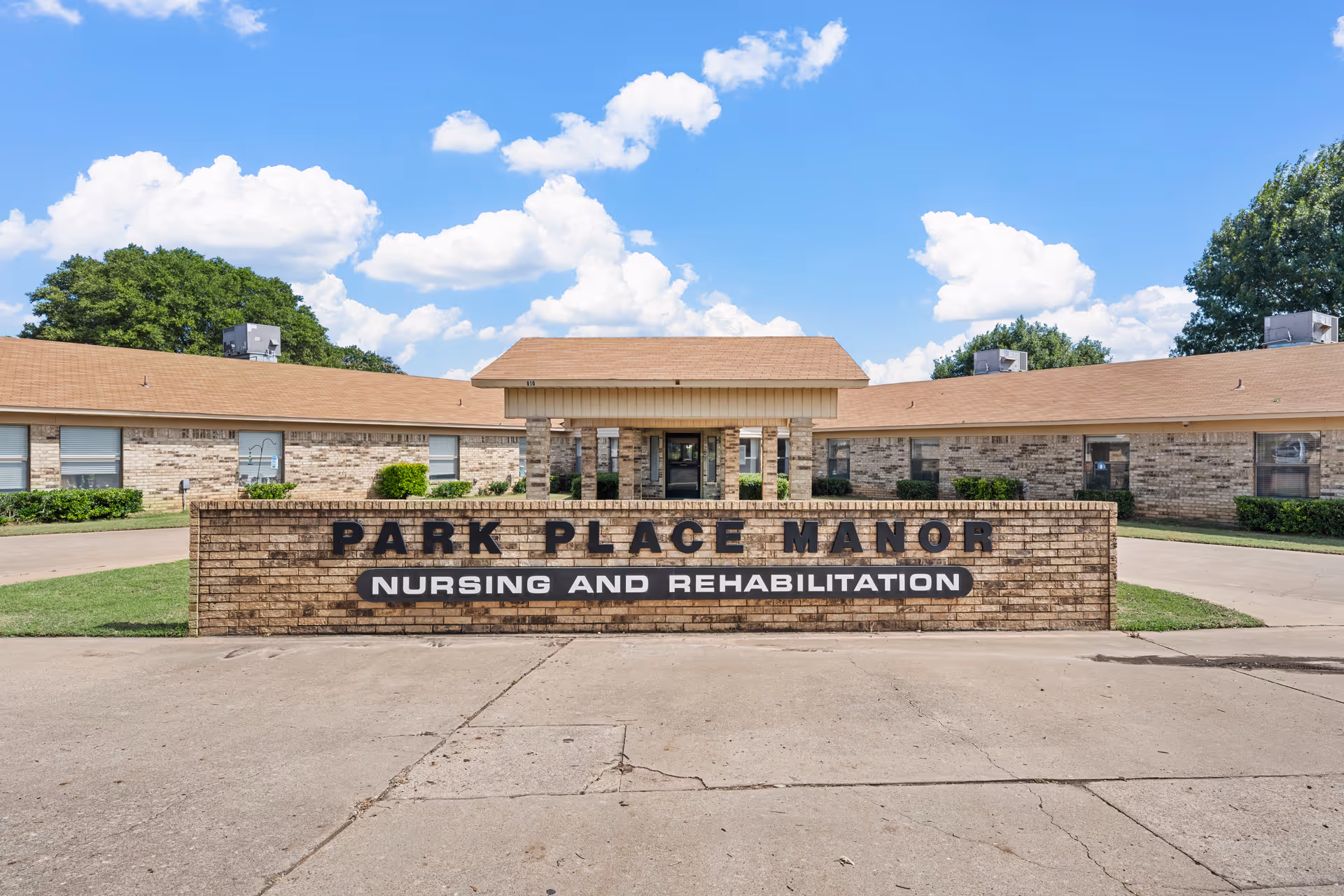 Front exterior view of Park Place Manor Nursing and Rehabilitation building with a brick sign in front, a driveway, and a clear blue sky with some clouds.