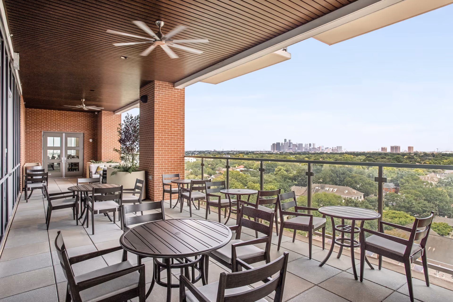 Outdoor covered patio area with multiple round tables and chairs arranged for seating. The patio has a wooden ceiling with ceiling fans and brick pillars. Beyond the glass railing, there is a view of a green landscape with trees and a distant city skyline under a clear sky.