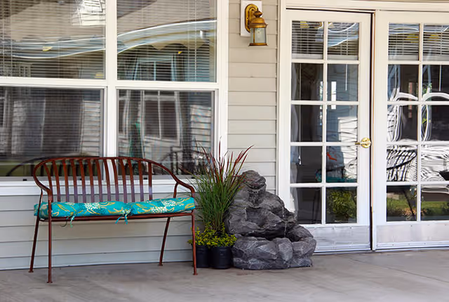 Outdoor patio area with a metal bench that has a green cushion, potted plants, a decorative rock water feature, and glass French doors leading inside a building.