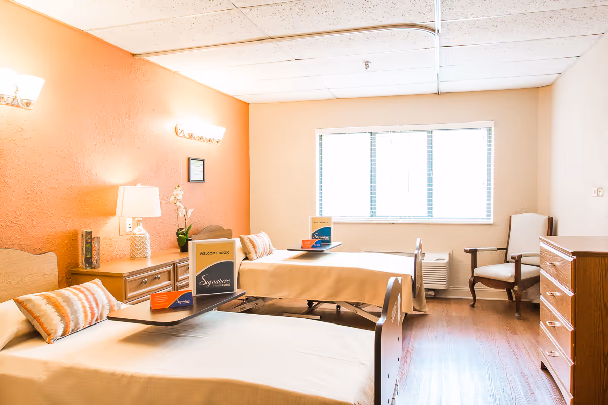 A bright and clean room with two single beds, each with a beige blanket and a decorative pillow. Between the beds is a wooden nightstand with a lamp, a small plant, and a welcome book labeled 'Signature HealthCARE.' There is a large window with blinds letting in natural light, a wooden dresser, and a cushioned armchair in the corner. The walls are painted in warm tones, with one wall in a soft orange color.