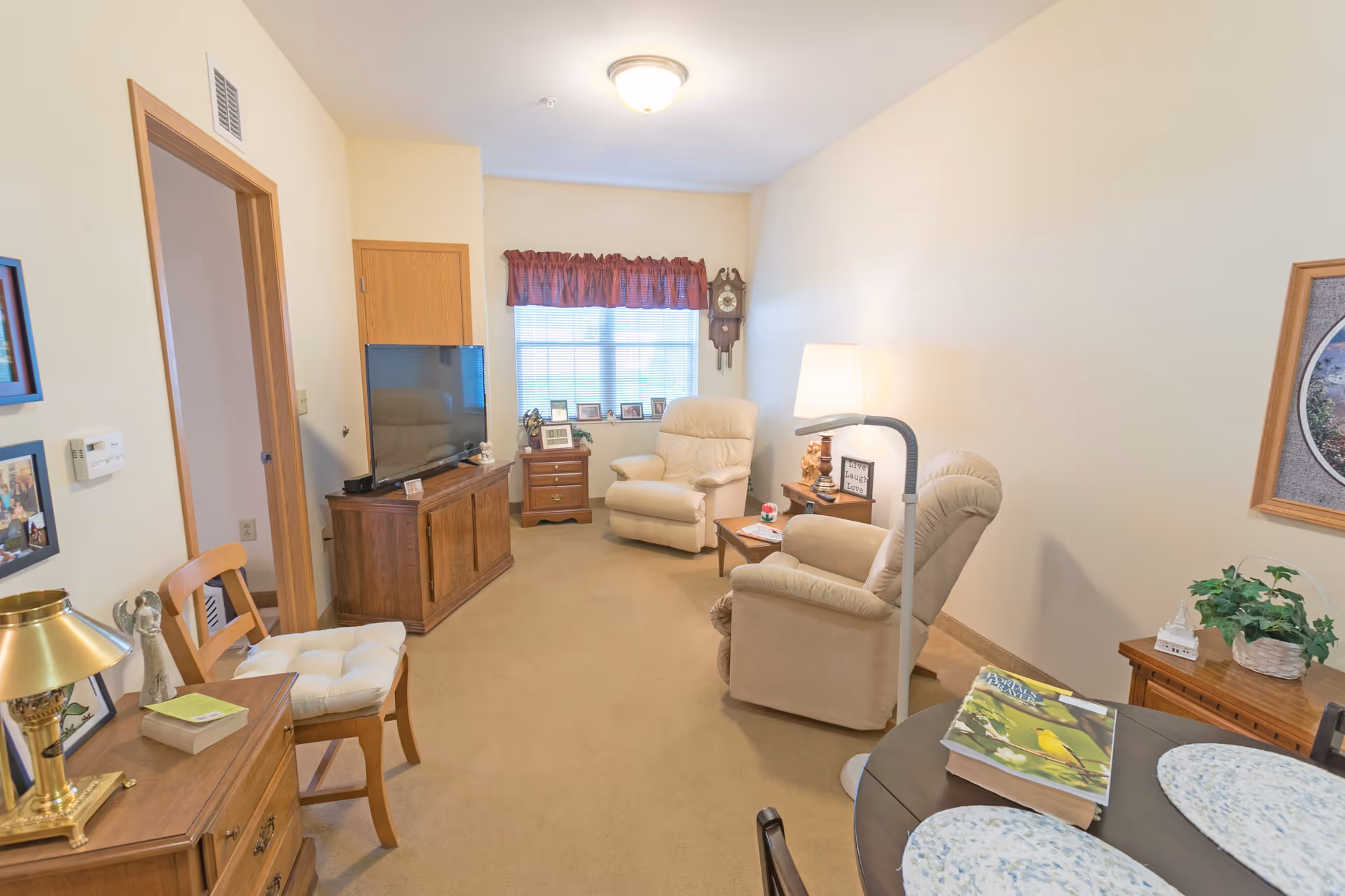 A cozy living room with beige walls and carpet, featuring two beige recliner chairs, a wooden TV stand with a flat-screen TV, a small wooden side table with a lamp, a round dining table with placemats and books, and a window with a red valance. Various framed photos and decorative items are placed around the room.