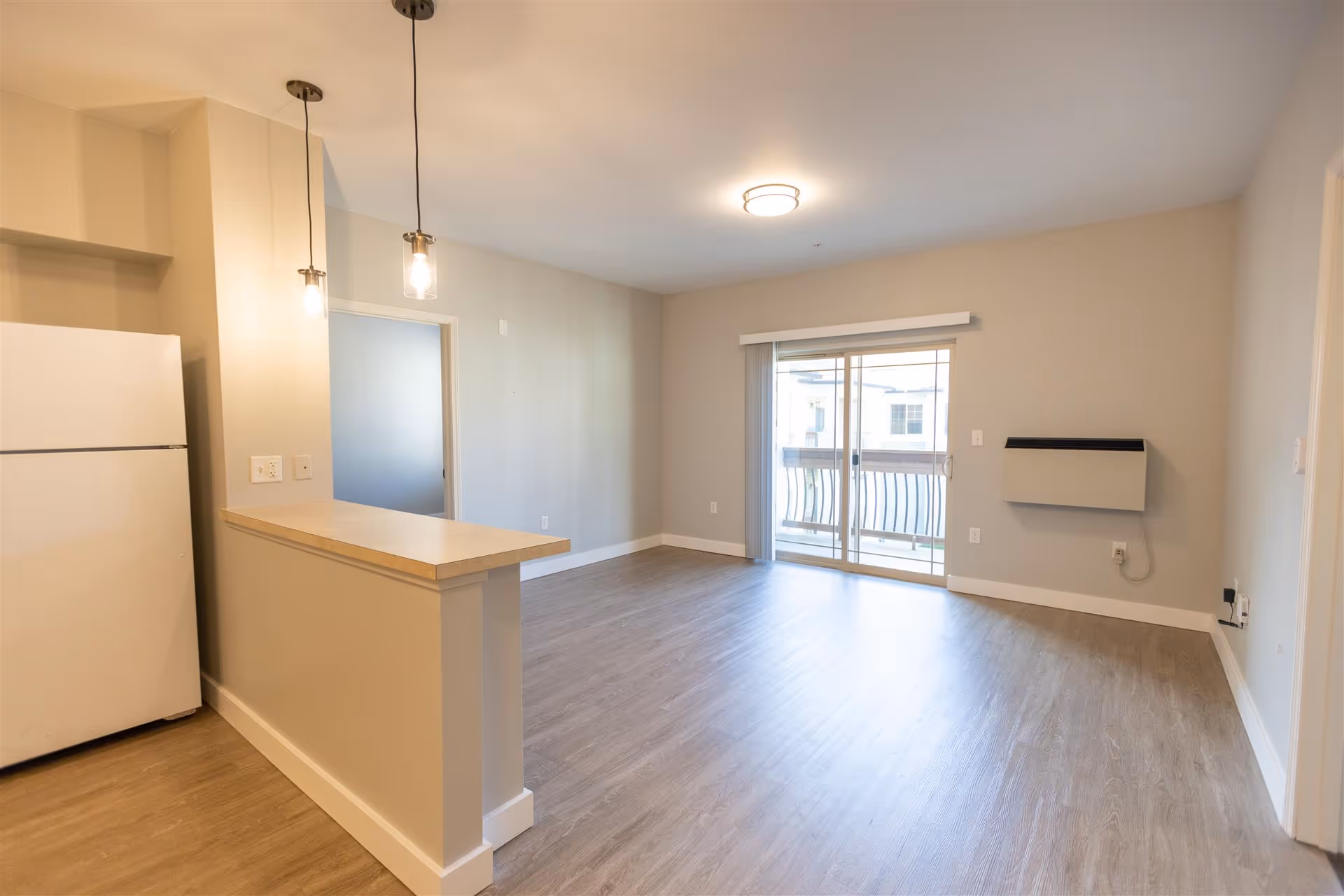 Empty open-concept apartment living area with wood-look floors, a small kitchen counter and refrigerator on the left, pendant lights, and sliding glass doors to a balcony.