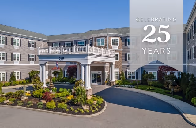 Front exterior view of The Bristal Assisted Living at West Babylon, showing a large building with multiple windows, a covered entrance with white columns, landscaped greenery, and a clear blue sky. A banner on the right side reads 'Celebrating 25 Years'.