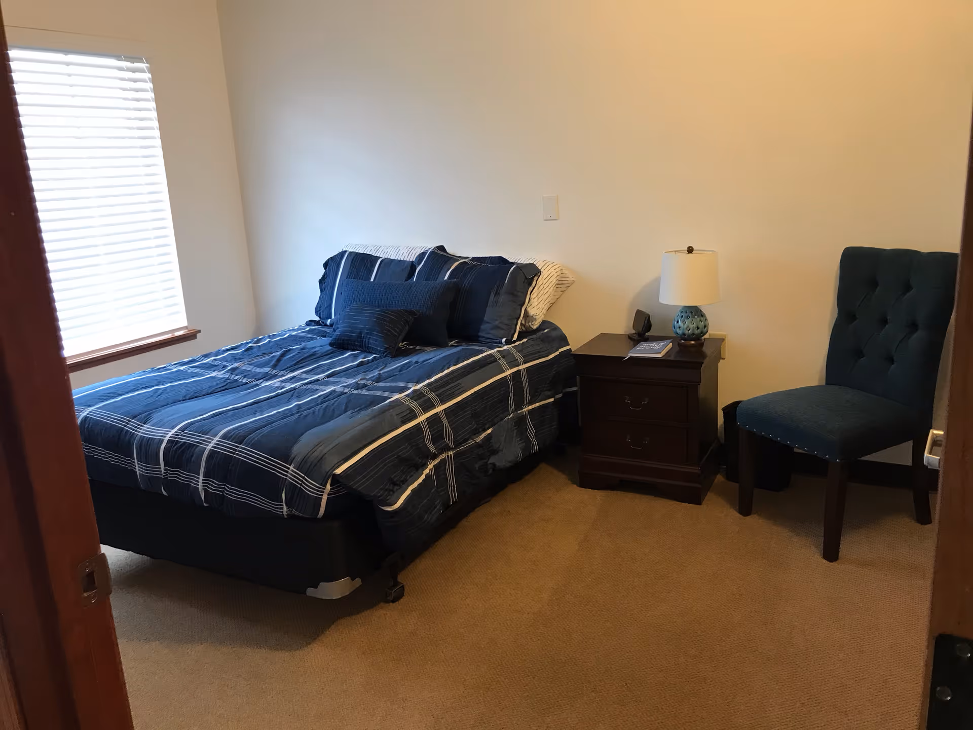 A small bedroom with a bed covered in blue and white striped bedding, a wooden nightstand with a lamp and a book, a blue upholstered chair, and a window with white blinds.