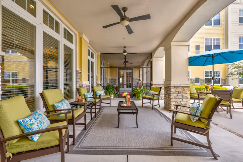 Covered outdoor patio area with green cushioned chairs and small tables arranged around a central coffee table on a large rug. Ceiling fans are mounted on the ceiling, and there are large windows and doors along one side. In the background, there is a blue patio umbrella and additional seating with green cushions.