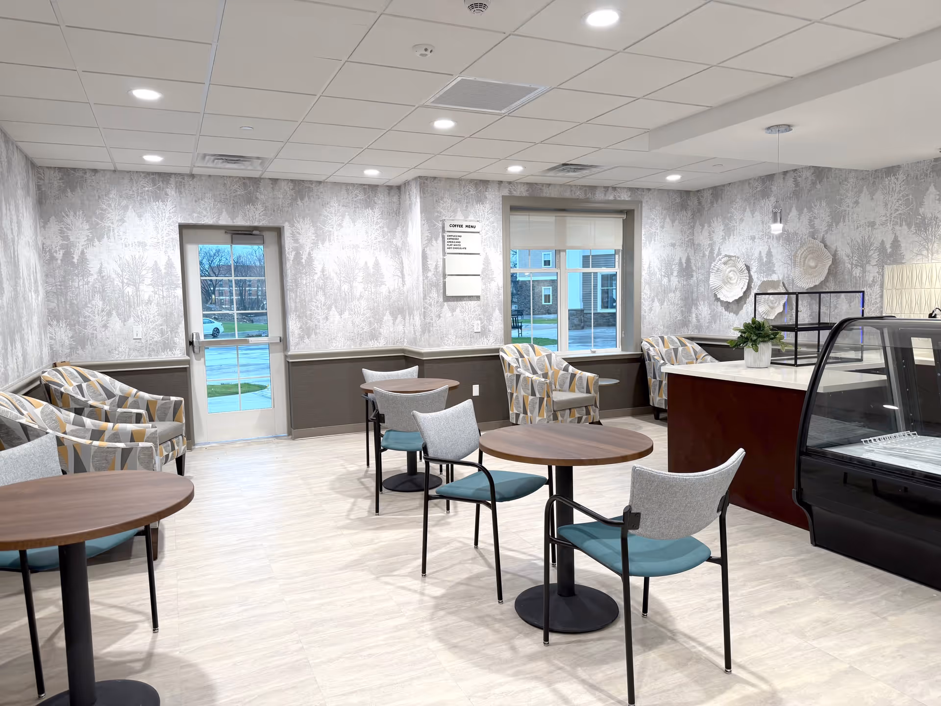 A bright and modern seating area with round wooden tables and chairs with teal cushions. There are patterned armchairs along the walls, a counter with a glass display case, and decorative wall art. The walls have a forest-themed wallpaper and large windows letting in natural light.