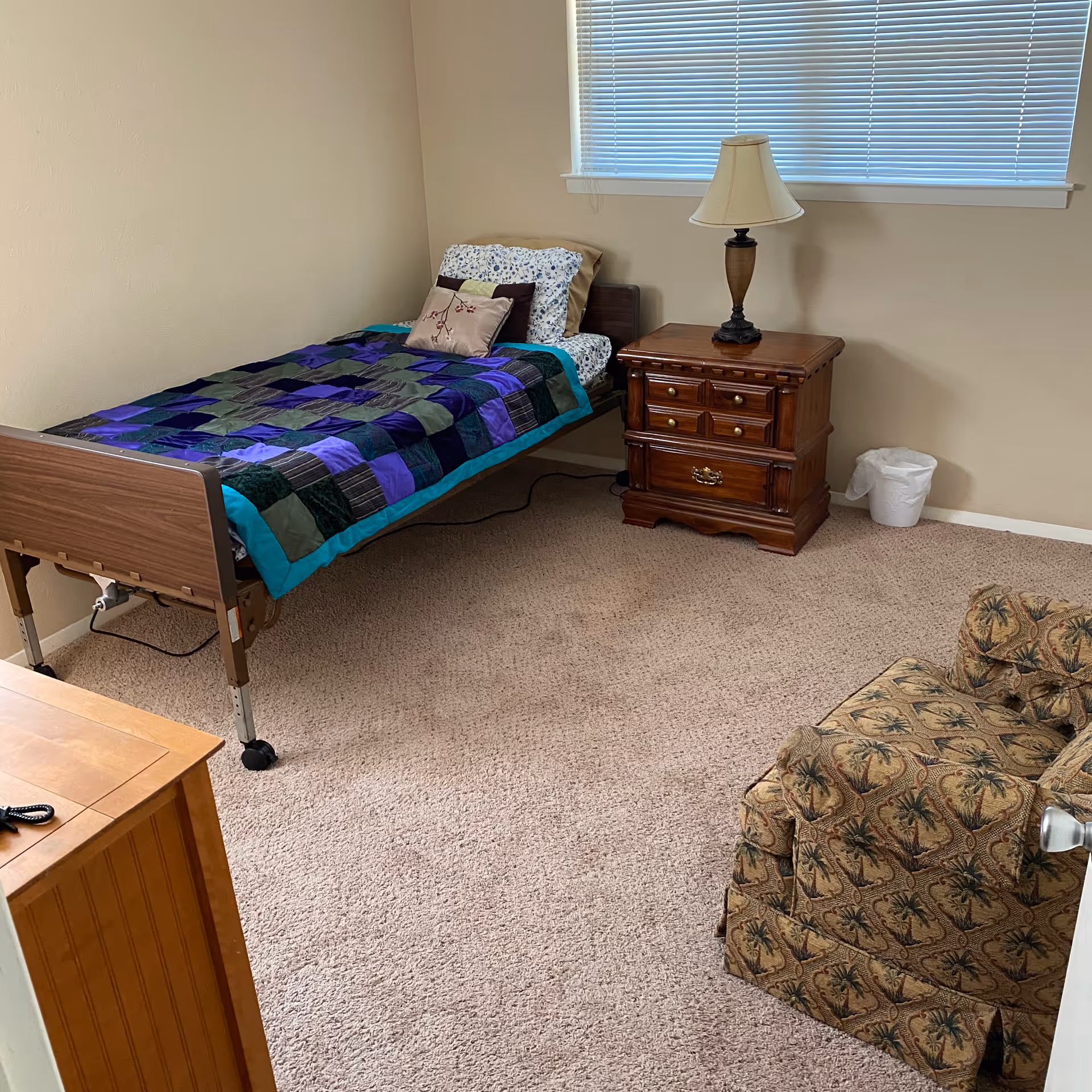 Carpeted senior bedroom with a single adjustable bed covered by a patchwork quilt, a wooden nightstand with a lamp, and a patterned armchair.