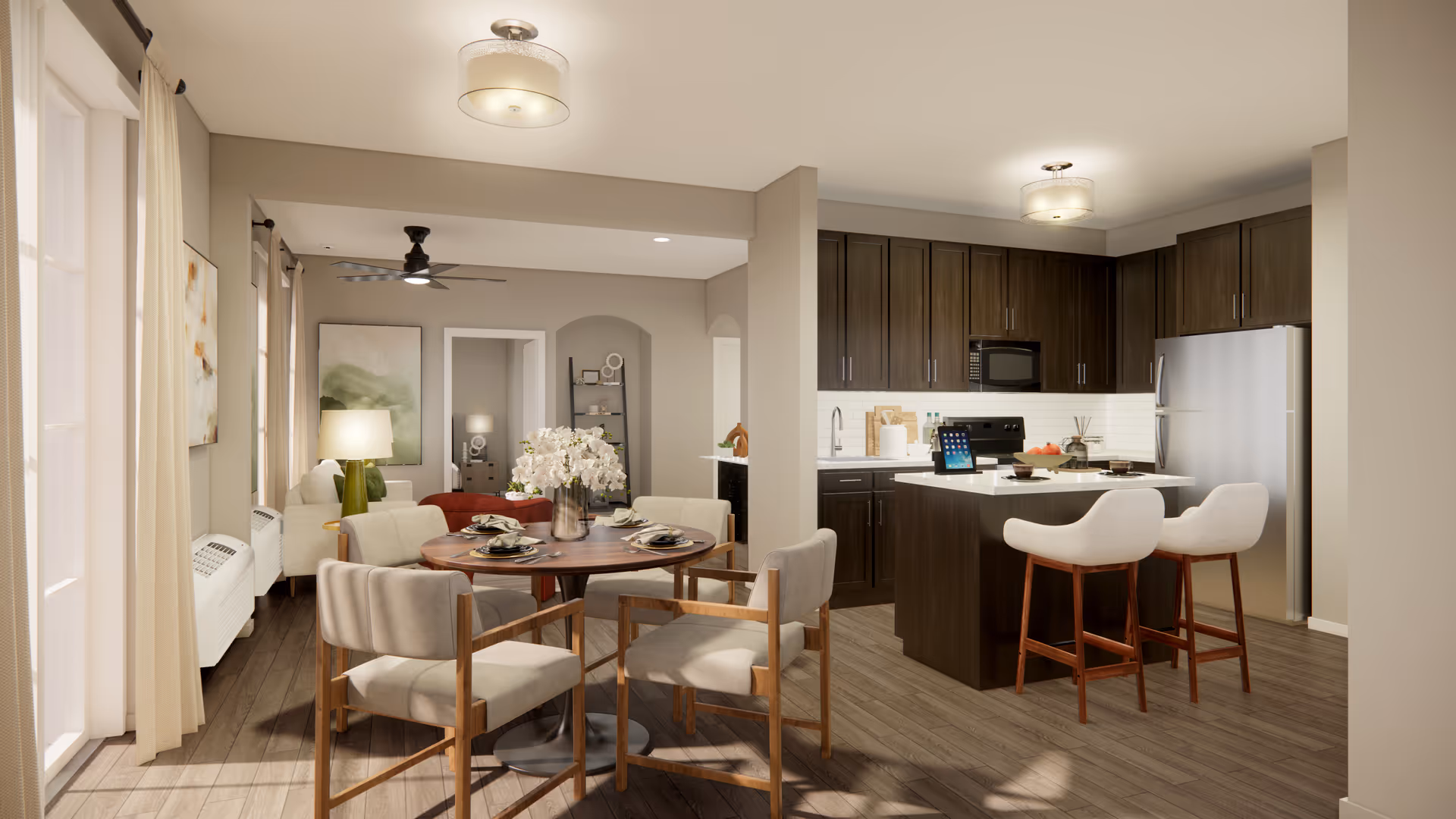 A bright and modern open-concept living space in a senior living facility featuring a kitchen with dark wood cabinets, a central island with two cushioned bar stools, and stainless steel appliances. Adjacent to the kitchen is a round dining table set with four chairs and a vase of white flowers. The living area includes a sofa, a floor lamp, and wall art, with large windows letting in natural light through beige curtains.