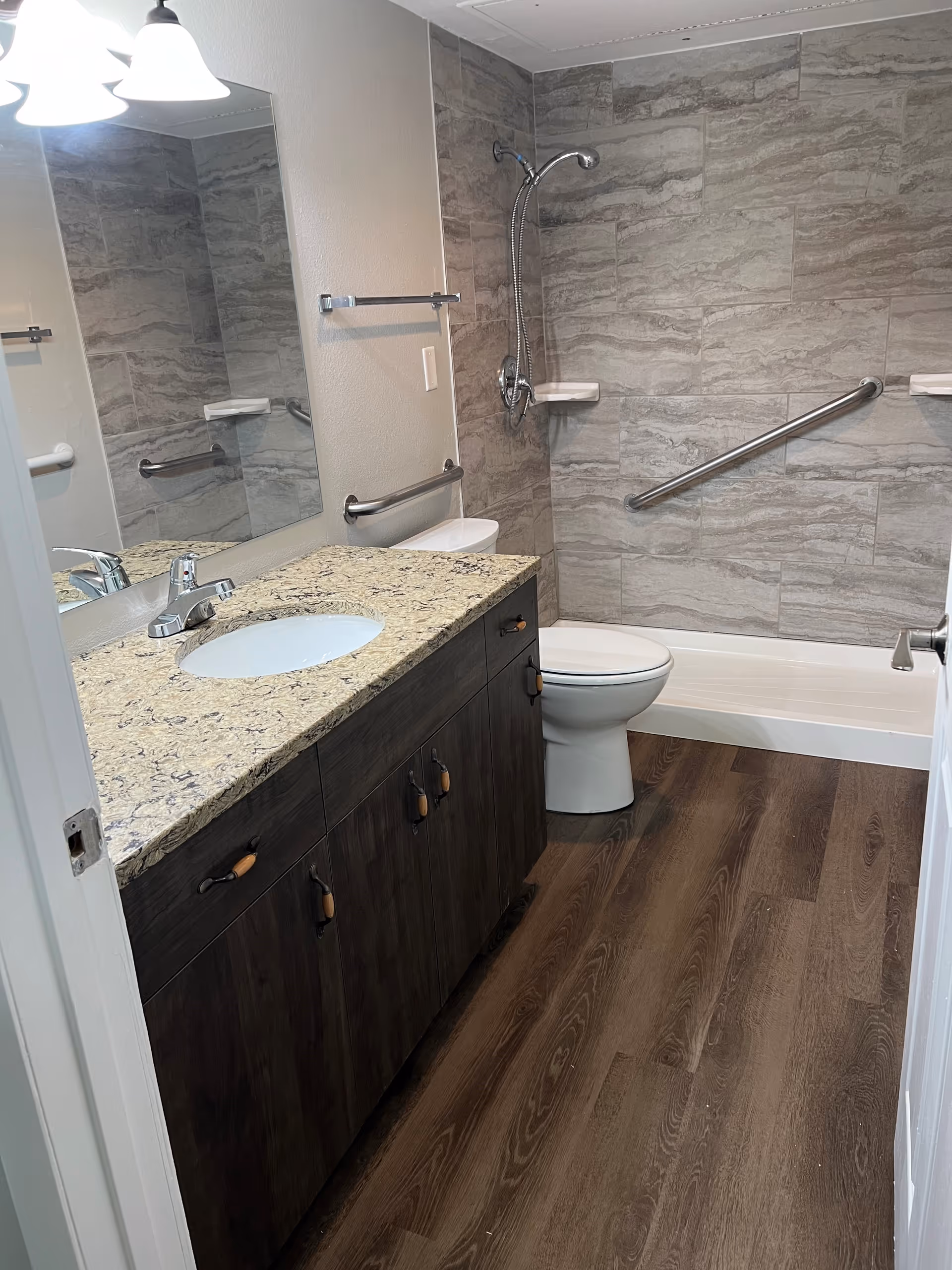 Accessible bathroom with granite counter, sink, mirror, toilet, and shower in a senior facility.