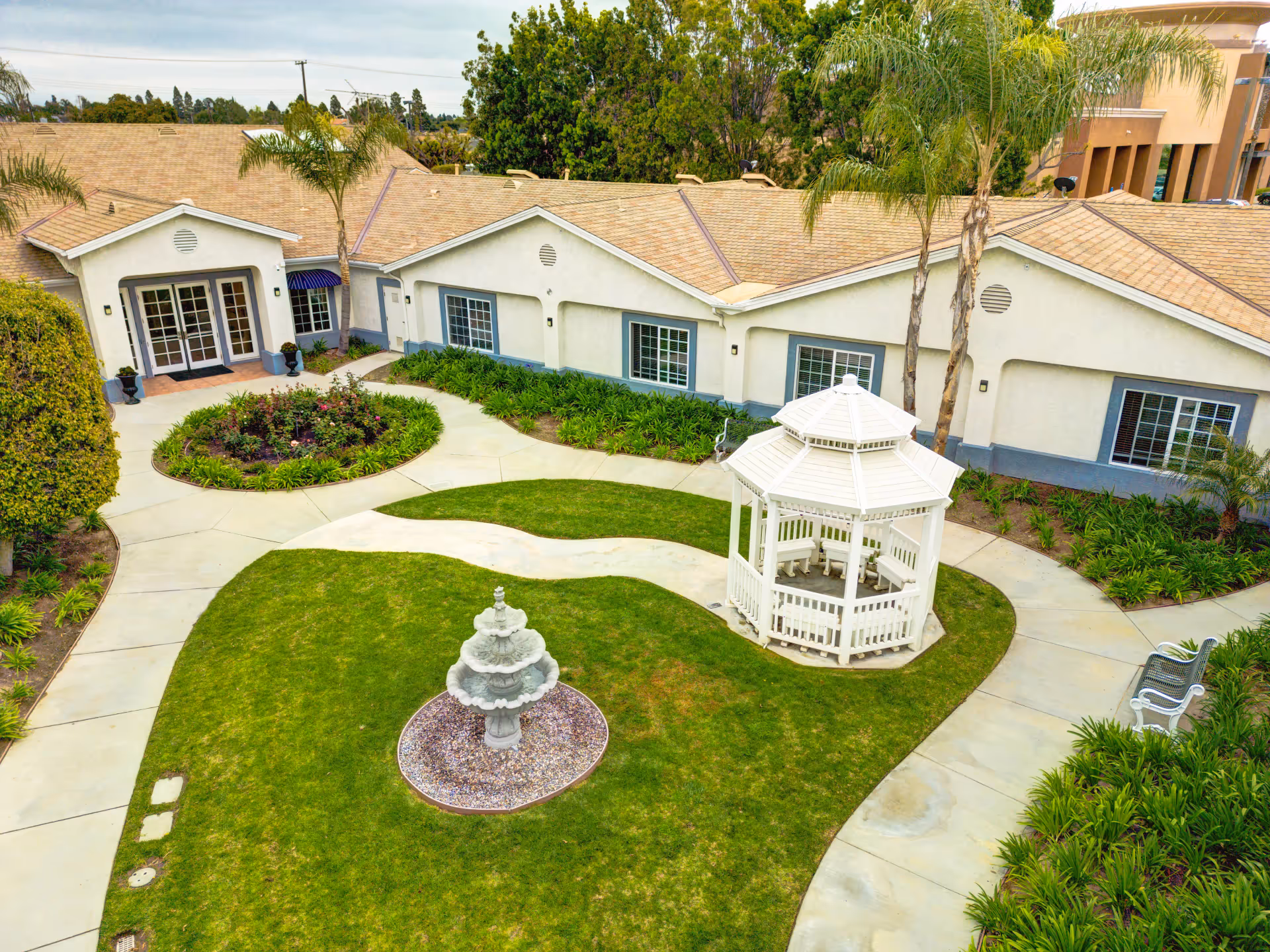 A well-maintained outdoor courtyard at Ivy Terrace at Garden Grove Memory Care featuring a white gazebo, a multi-tiered stone fountain, green grass, landscaped plants, and a paved walkway surrounding the area. The building with beige walls and multiple windows is visible in the background.