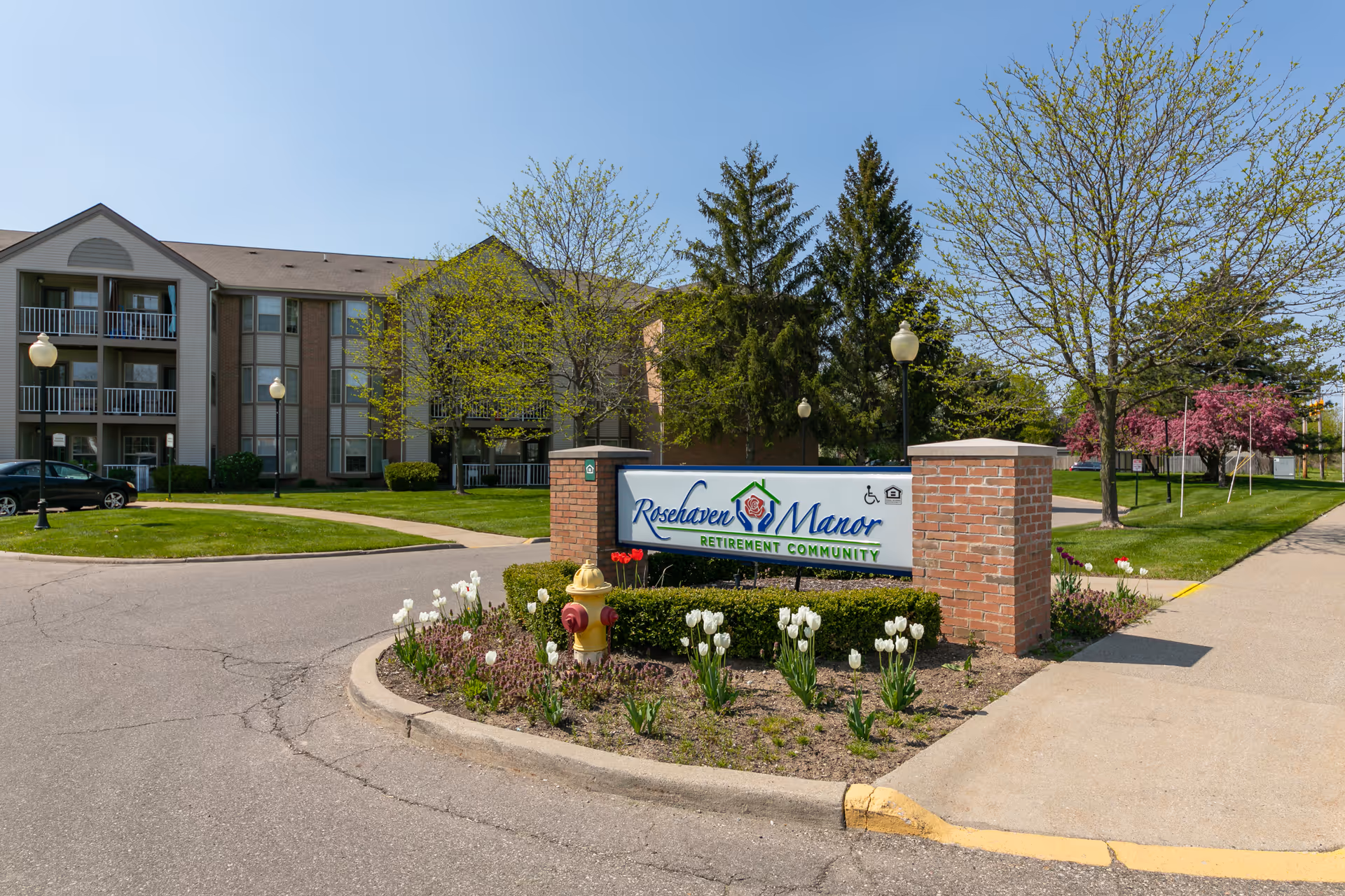 Entrance sign for Rosehaven Manor Retirement Community with a brick base surrounded by white tulips and a fire hydrant. Behind the sign is a multi-story residential building with balconies, green lawns, trees, and street lamps under a clear blue sky.