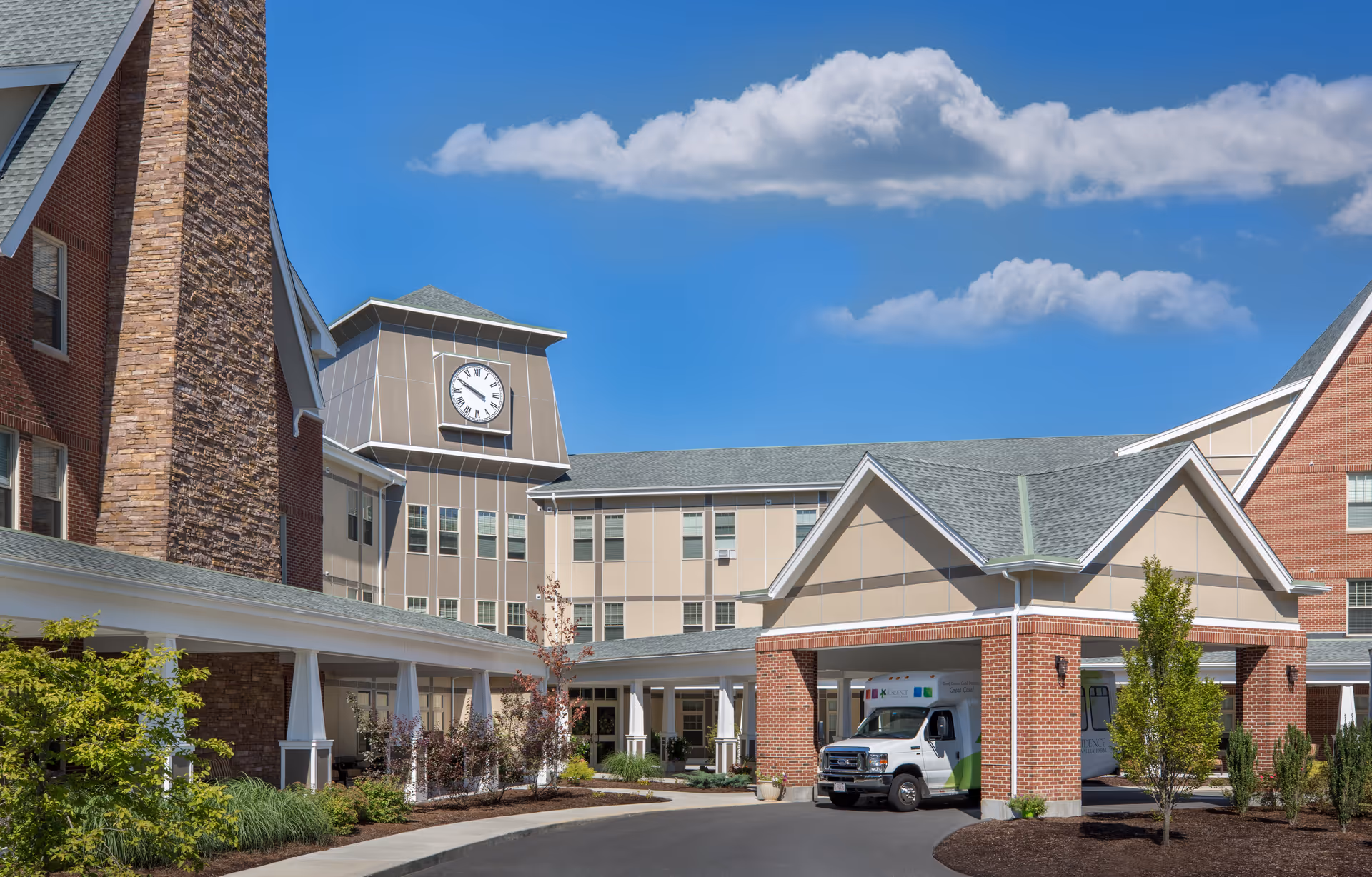 Exterior view of The Residence at Valley Farm senior living facility showing a multi-story building with a clock tower, brick and beige siding, a covered entrance with a vehicle parked underneath, landscaped greenery, and a clear blue sky with some clouds.