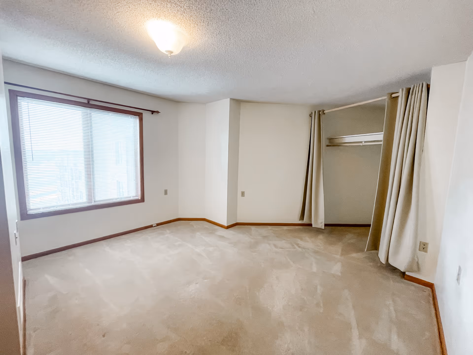 Empty carpeted bedroom with a large window, overhead light, and an open closet concealed by curtains.