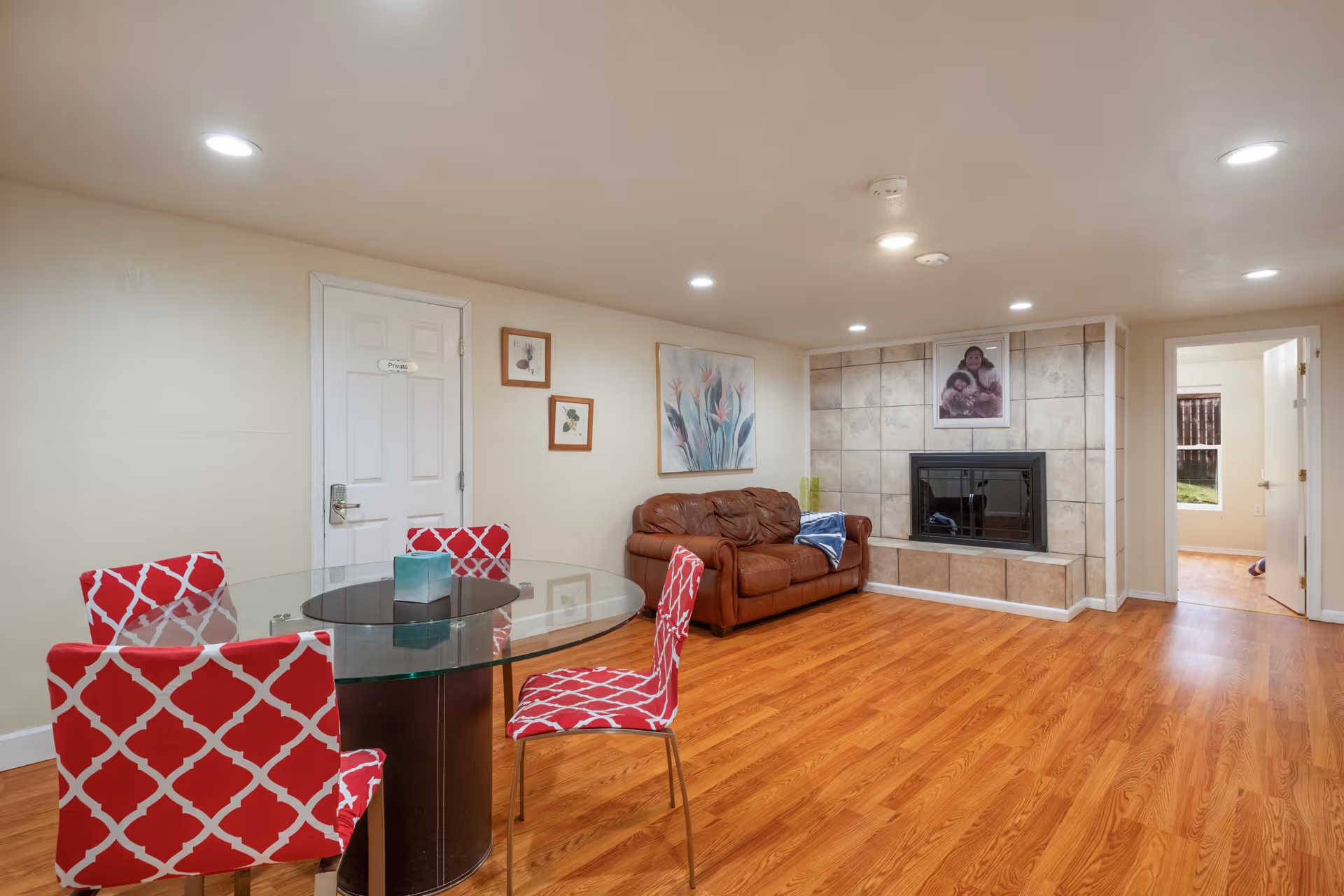 A cozy living room with wooden flooring, a brown leather couch, a glass round dining table with four red patterned chairs, and a tiled fireplace with a framed photo above it. The walls are light-colored with some framed artwork, and there are recessed ceiling lights. A door labeled 'Private' and an open doorway leading to another room are visible.