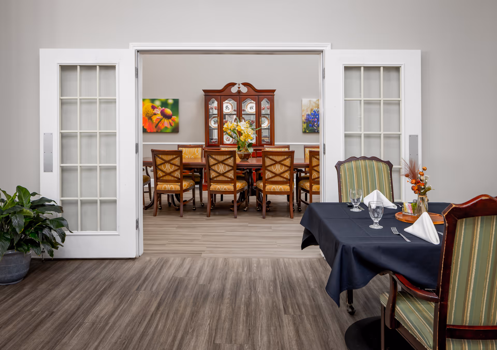 View of a dining area in a senior living facility with a table set with glasses, napkins, and a small floral centerpiece in the foreground. Beyond a set of white French doors, there is a larger dining table with chairs and a wooden china cabinet displaying plates. The walls are decorated with colorful floral paintings.