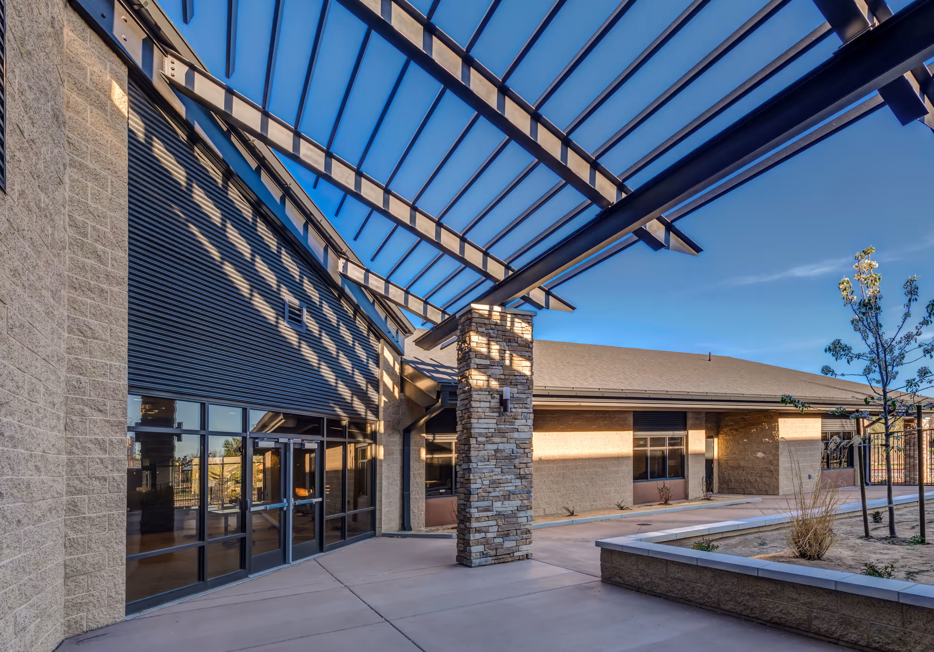 Outdoor view of a modern building with stone and brick walls, large glass doors, and a metal pergola casting shadows on the ground. There is a small landscaped area with young trees and plants under a clear blue sky.
