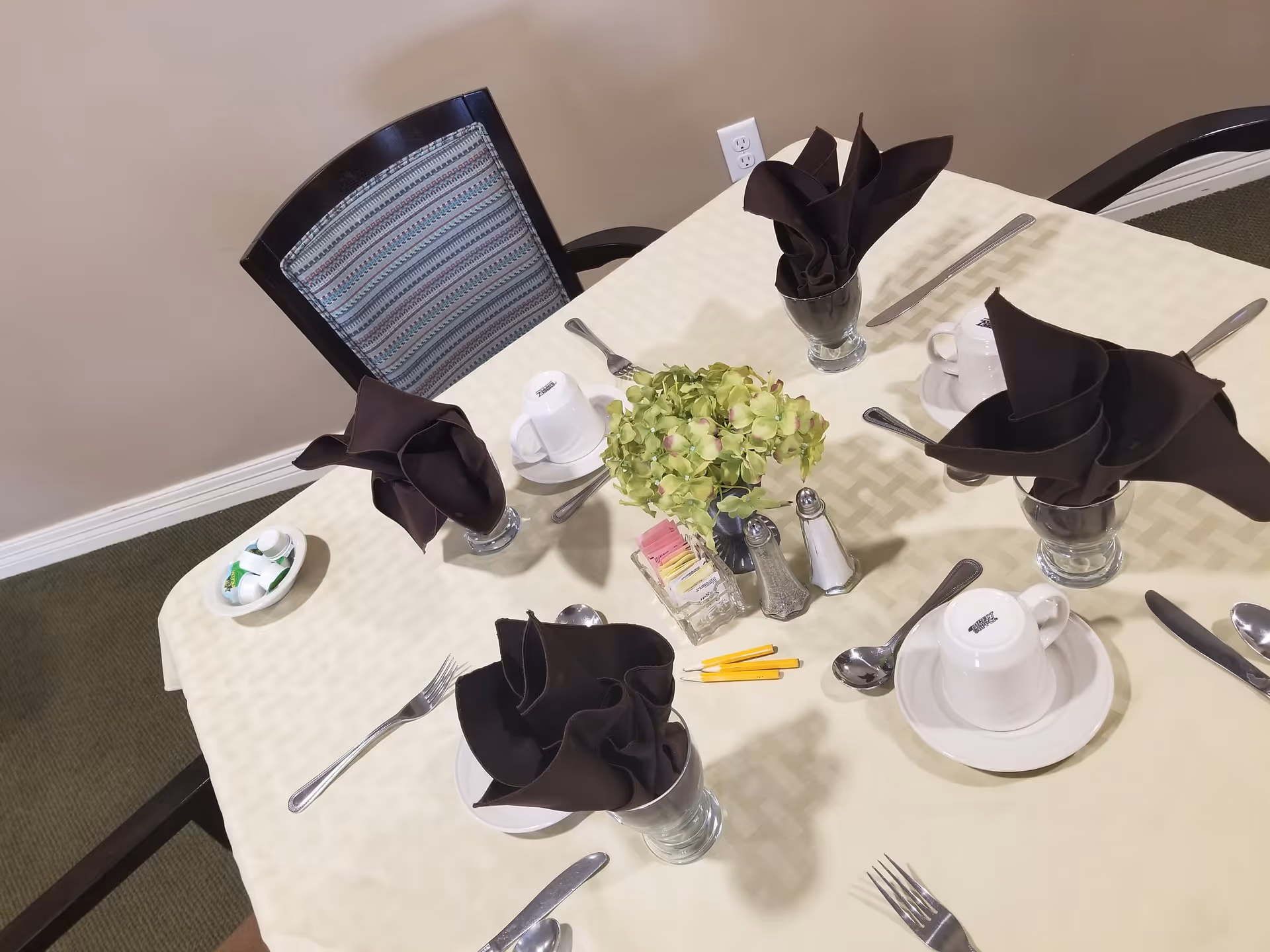 A dining table set for four with a cream-colored tablecloth, black folded napkins in glasses, white cups and saucers, silverware, a small bowl with creamers, a centerpiece of green flowers, salt and pepper shakers, and packets of sugar and sweeteners. A chair with a patterned backrest is visible at the table.