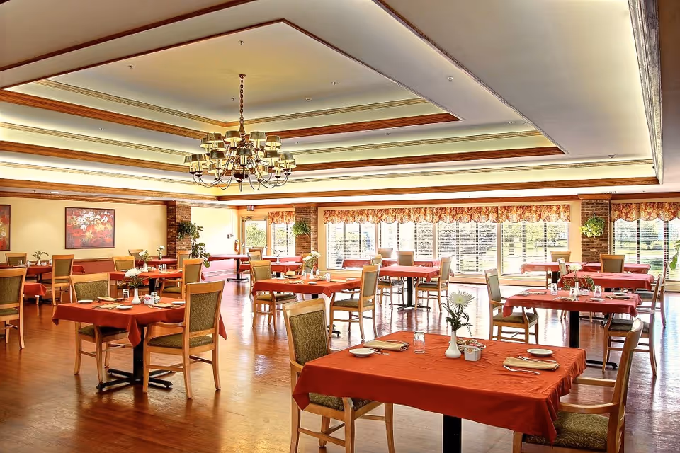 Spacious dining room with multiple tables covered in red tablecloths, wooden chairs, a central chandelier, and large windows letting in natural light.