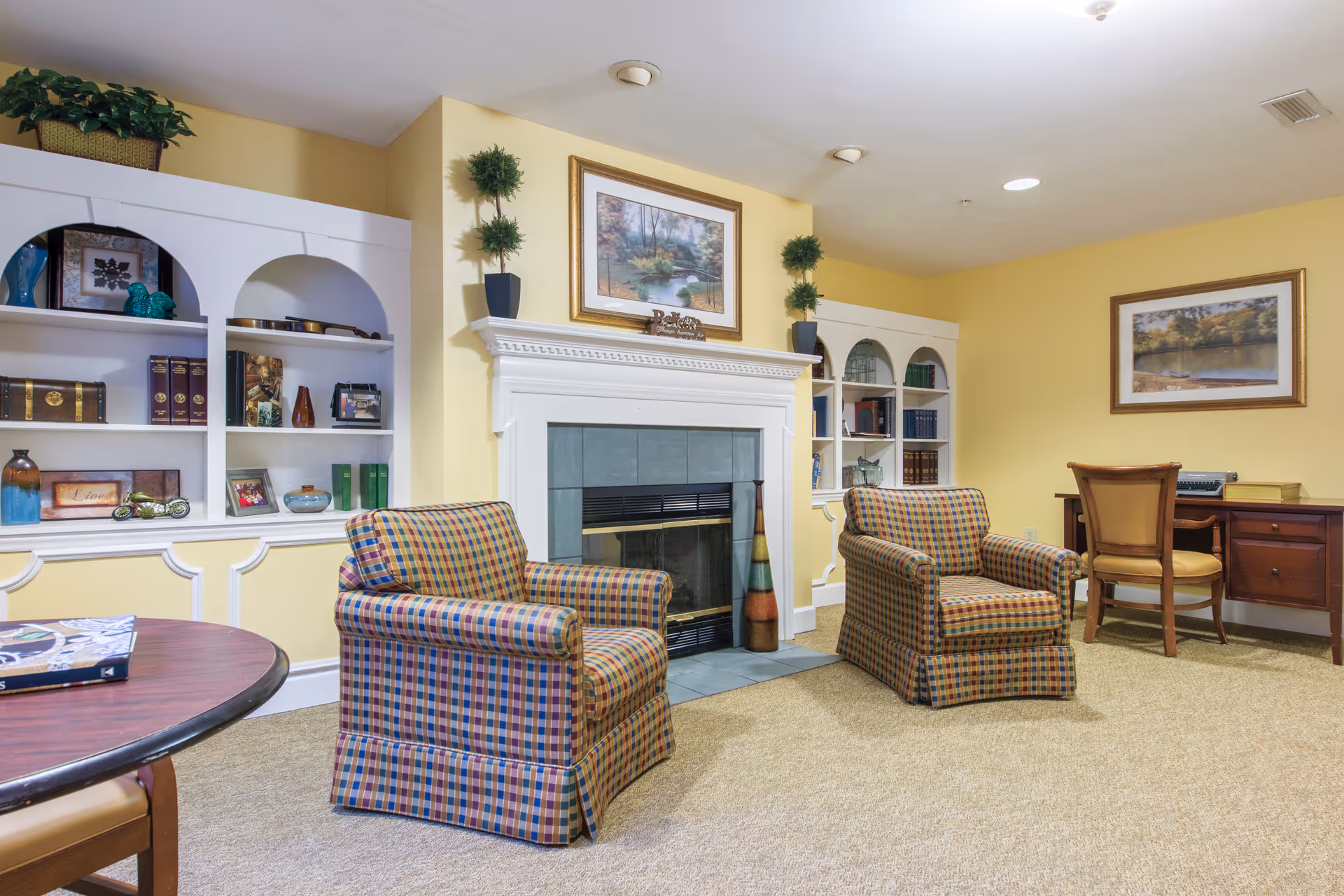 Cozy sitting room with two plaid armchairs facing a fireplace, built-in shelving, and a desk.