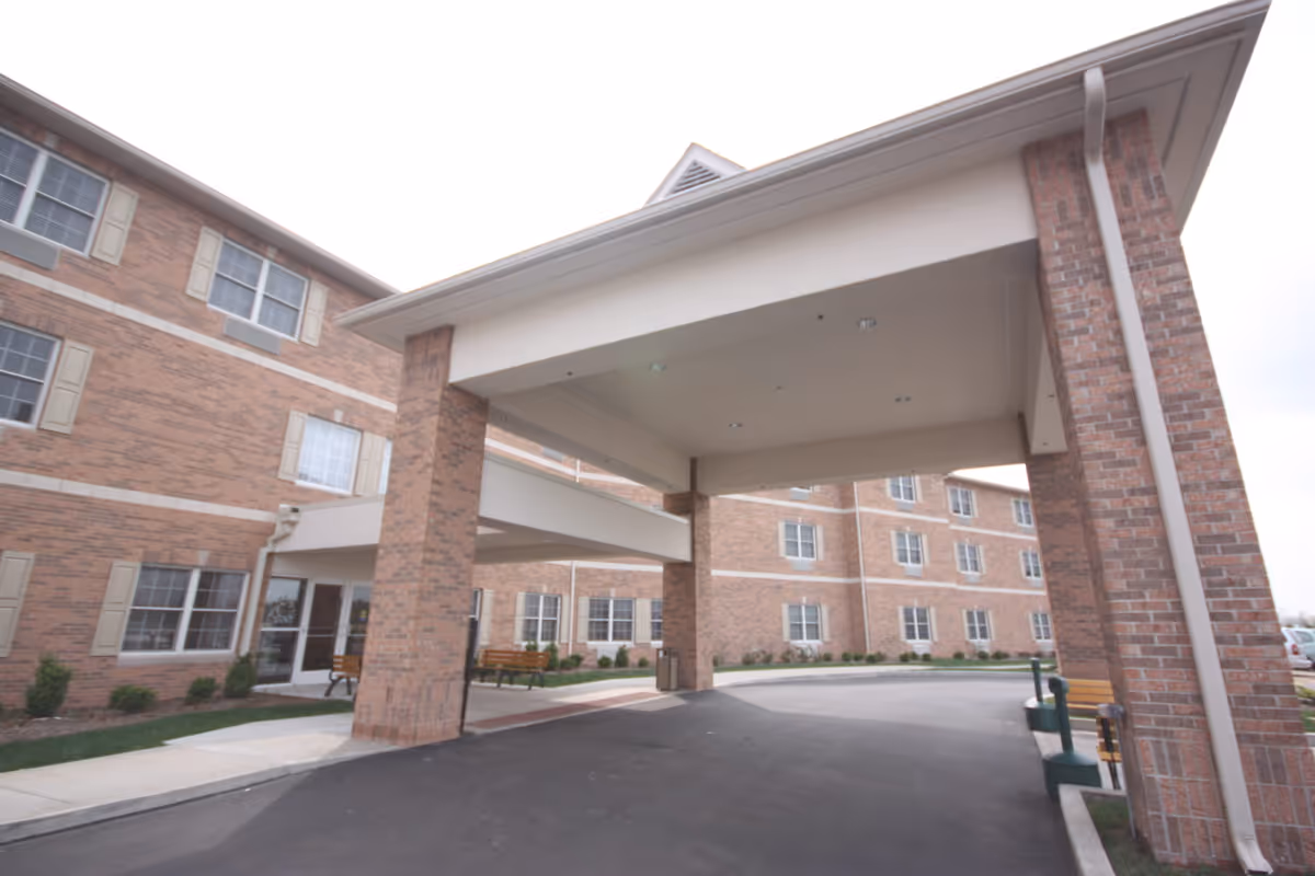 Exterior view of a multi-story brick building with a covered entrance supported by large brick pillars, benches along the sidewalk, and windows with beige shutters.